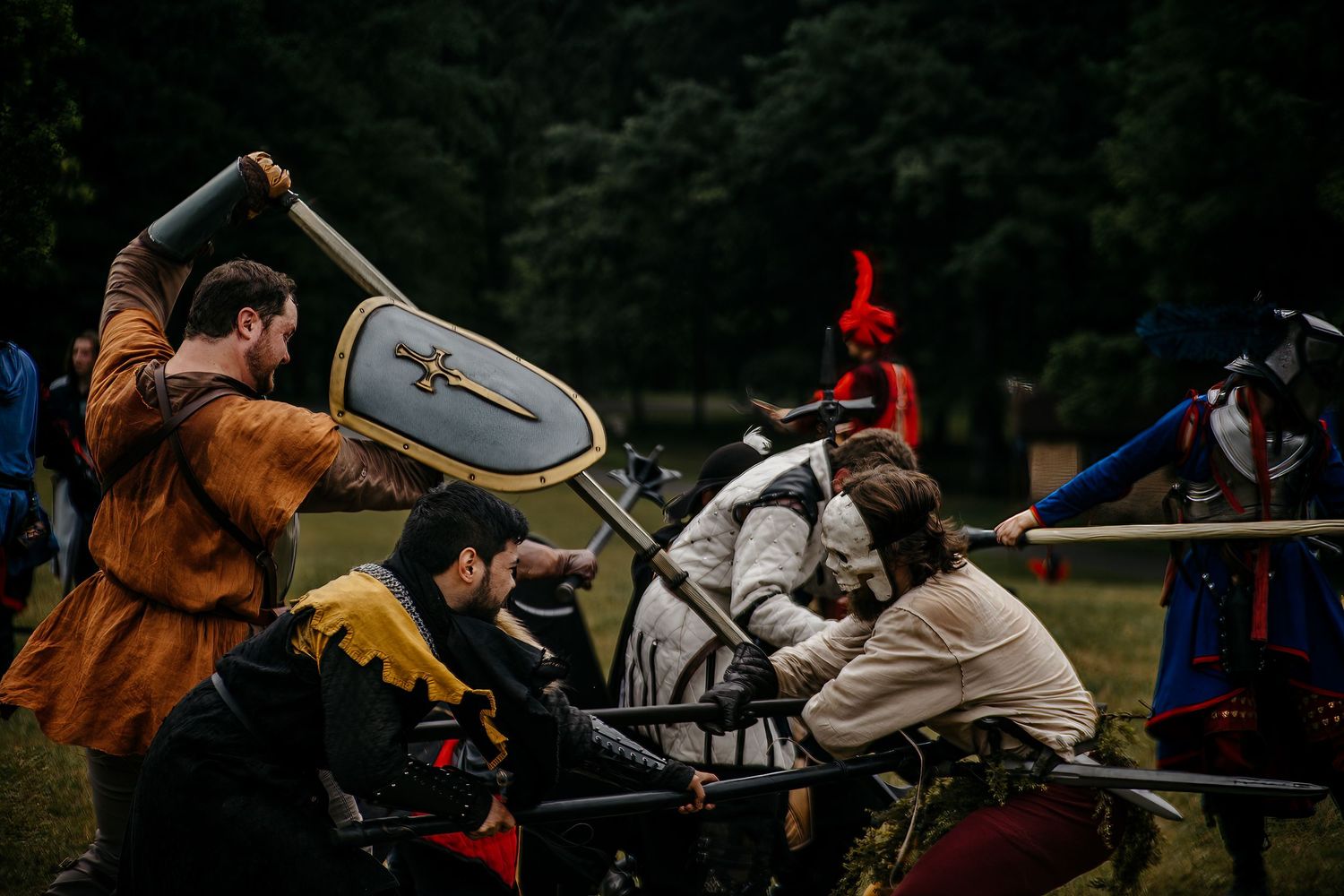 Medieval reenactment battle scene with knights in armor wielding shields and swords in an outdoor field.