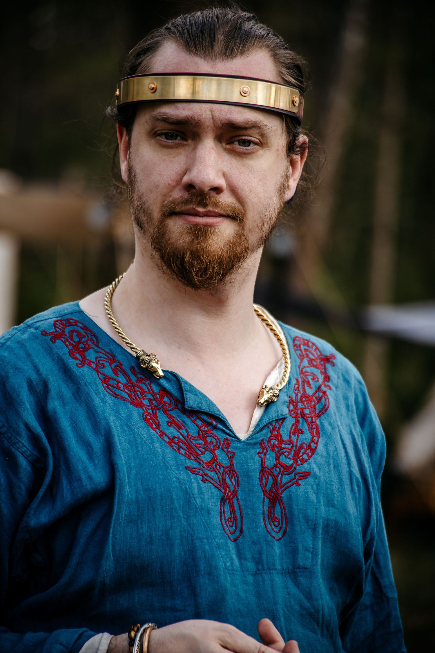 A bearded man wearing a teal embroidered tunic, gold headband, and chain necklace in a medieval Viking costume.