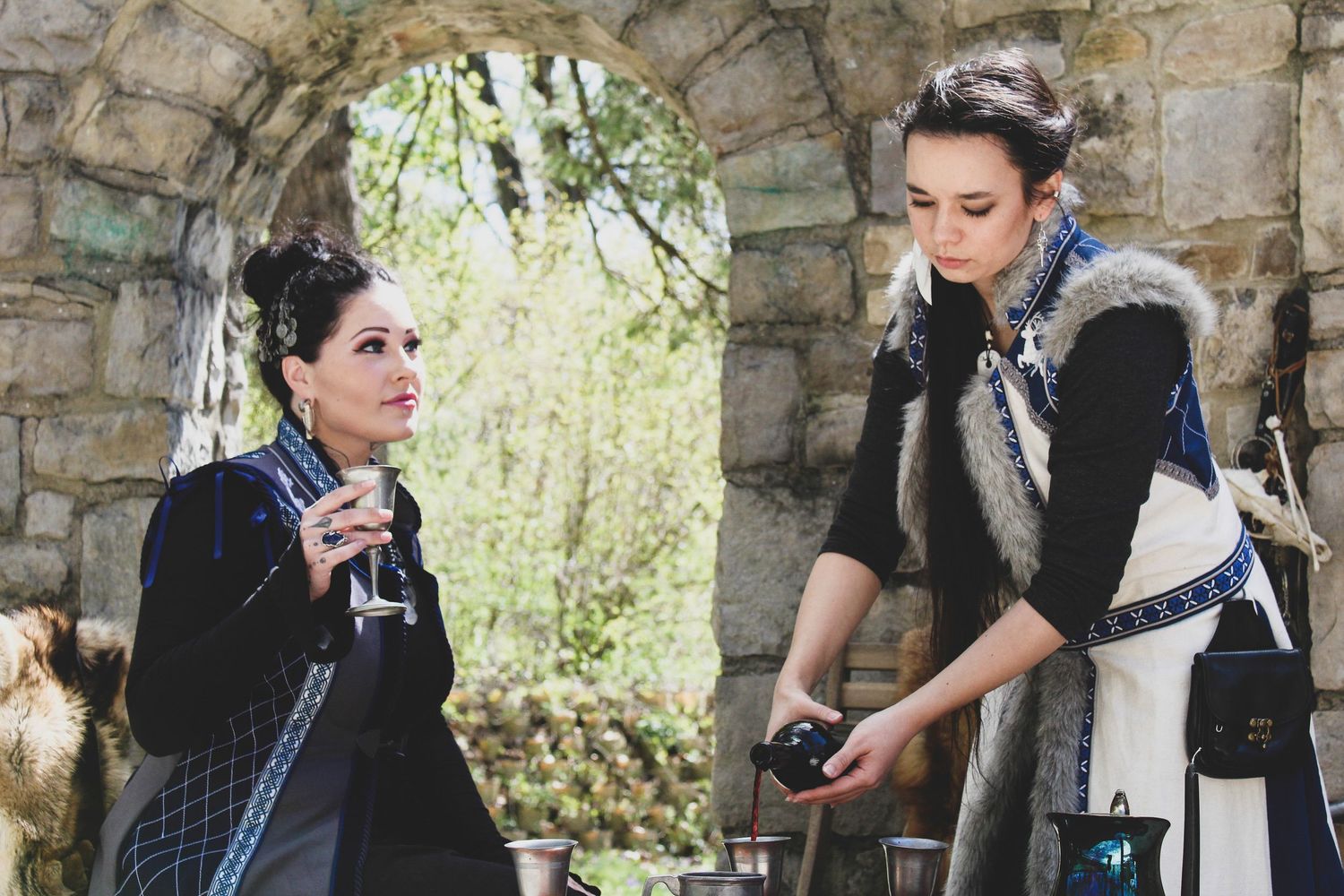 Two elegantly dressed women in medieval-inspired fashion converse outdoors near a stone archway.