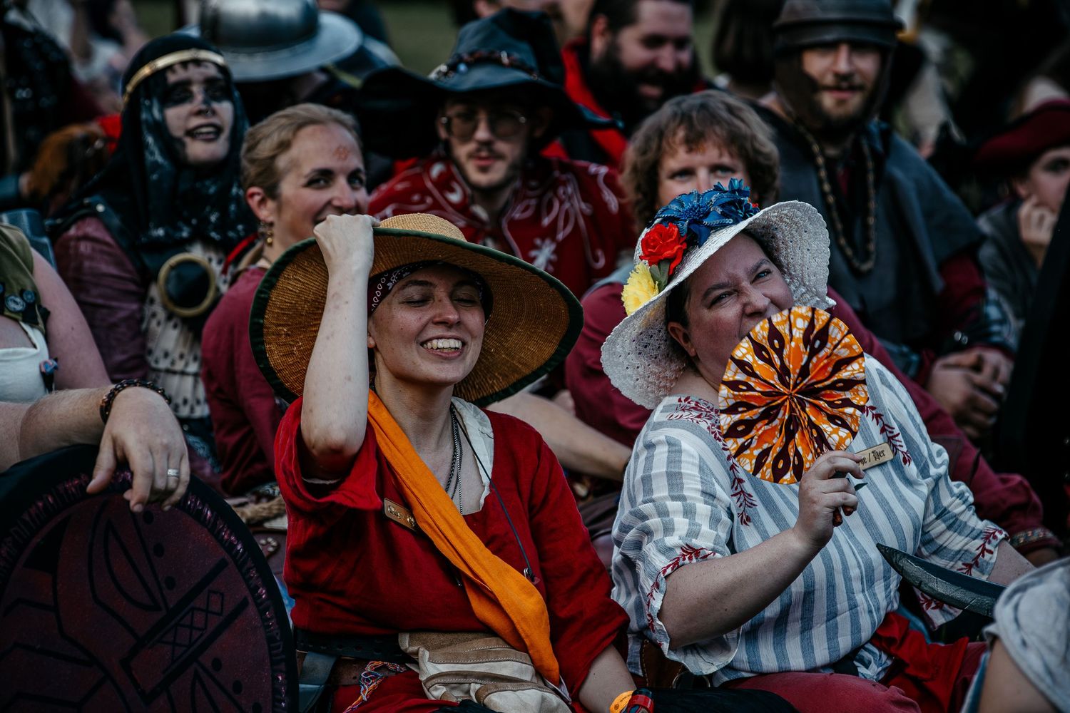 Costumed festival-goers in Renaissance faire attire smile and hold decorative fans at an outdoor event.