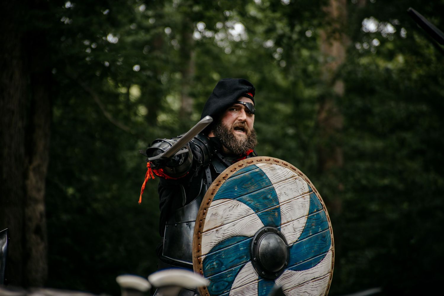 Bearded man in medieval Viking armor holding a blue shield in a forest battle reenactment.