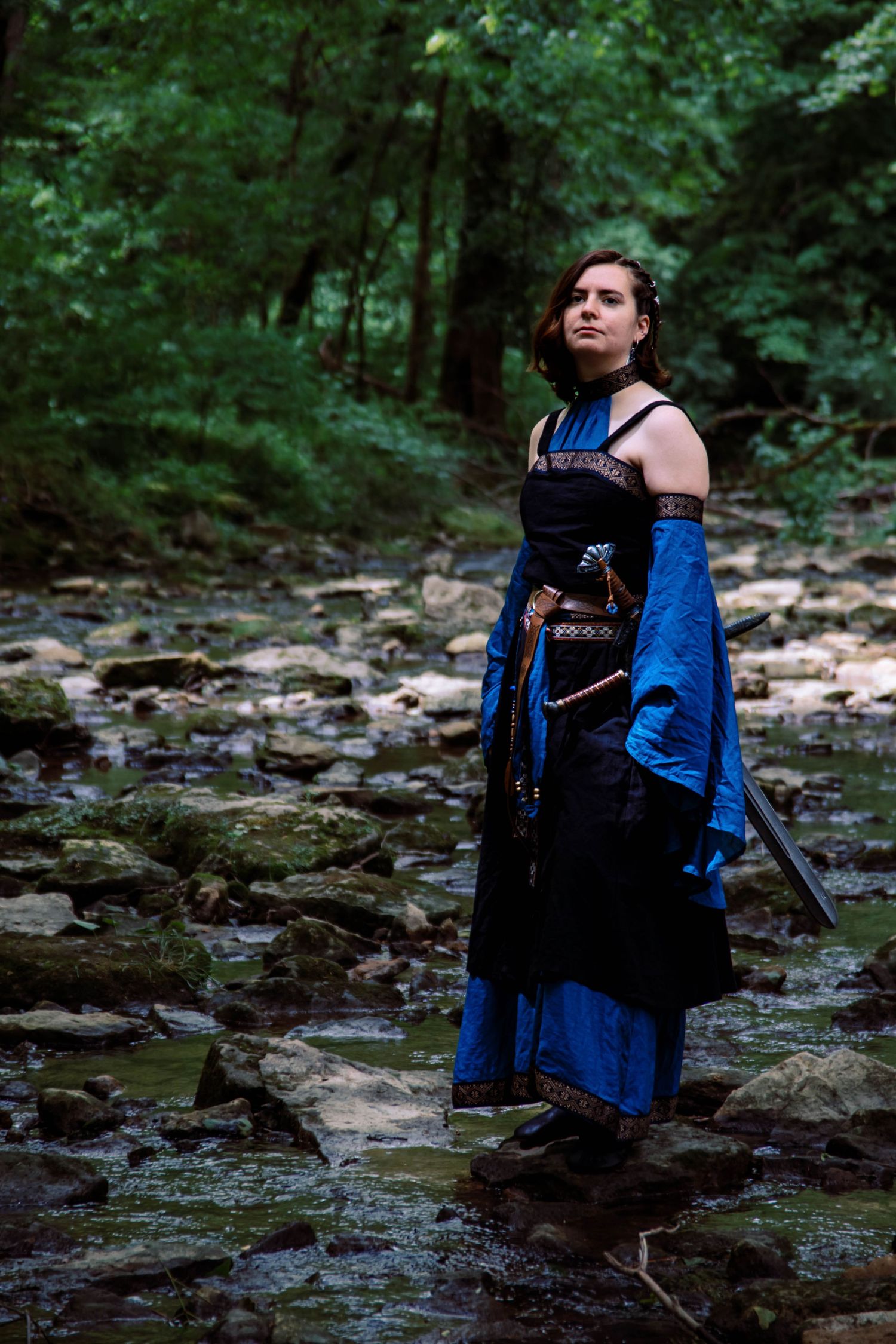 A figure in a flowing blue dress stands among rocky streams in a mystical forest setting.