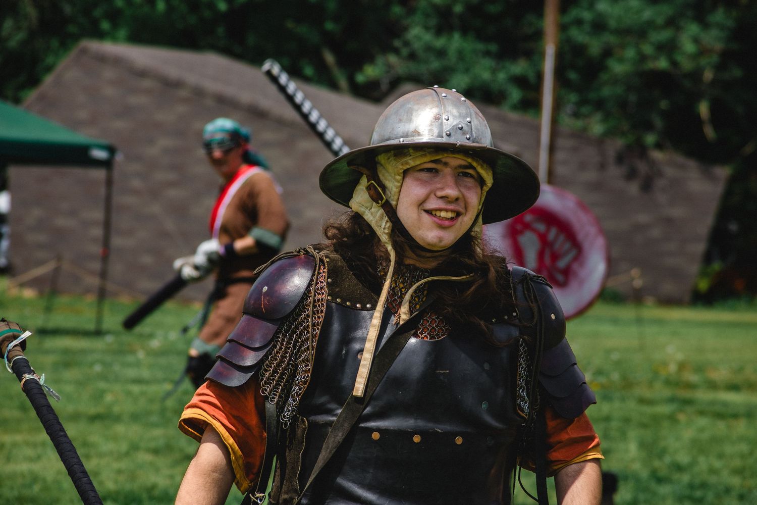 Young warrior in medieval armor and helmet smiles at Renaissance faire event outdoors.