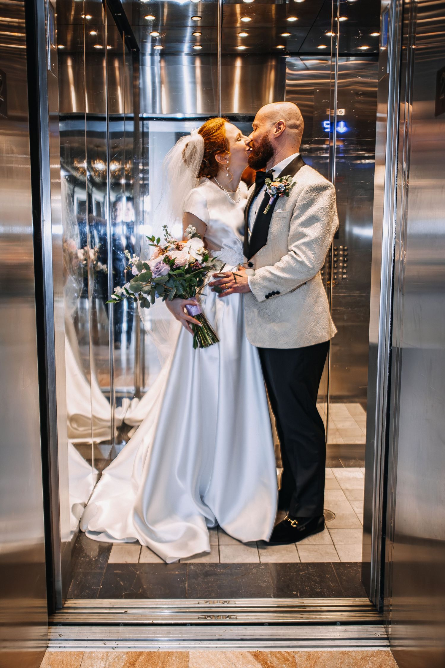 Bride and groom sharing a kiss in a mirrored elevator, bride holding a floral bouquet in a white gown.