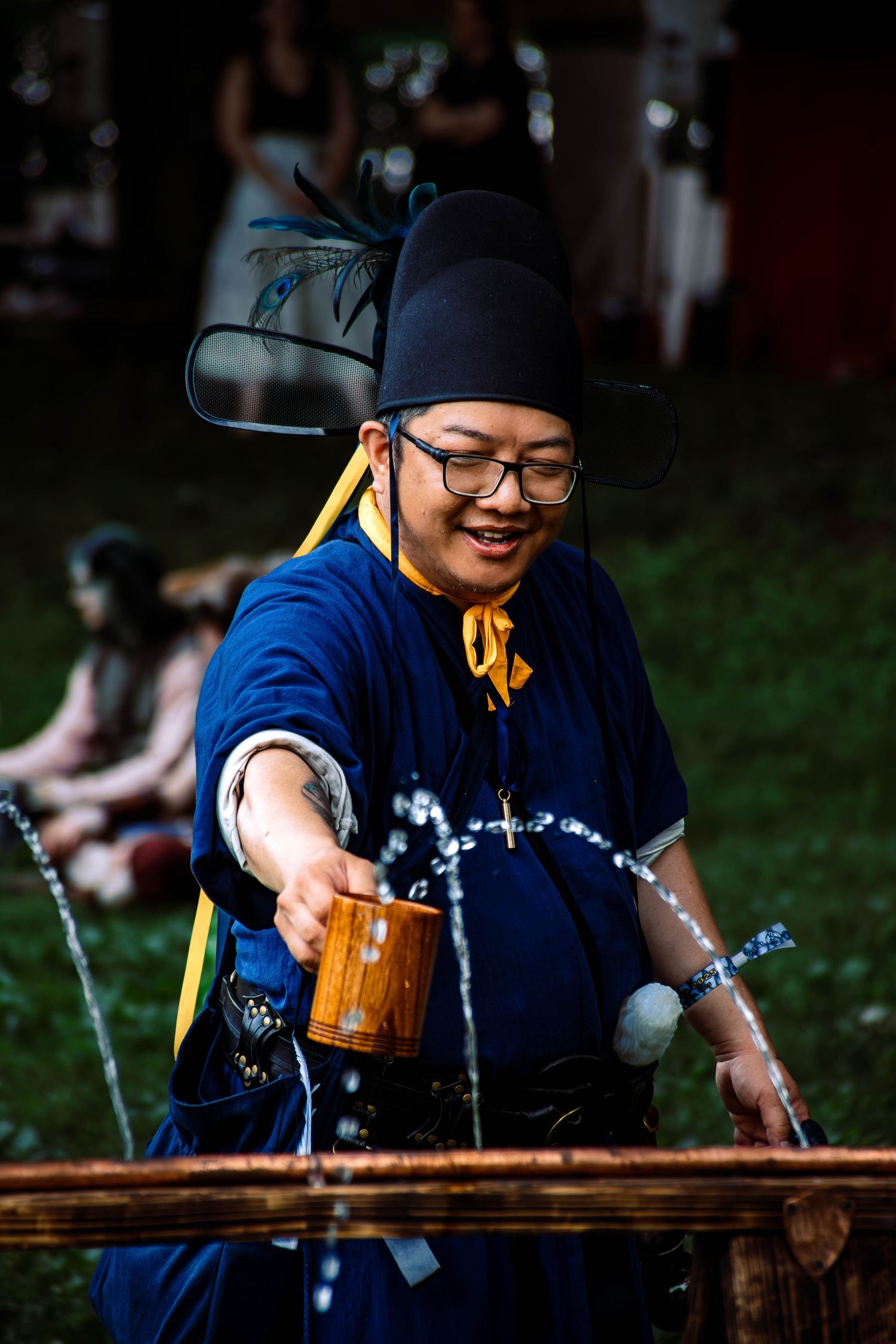 Someone wearing glasses and a blue uniform pours drinks at a wooden counter outdoors.
