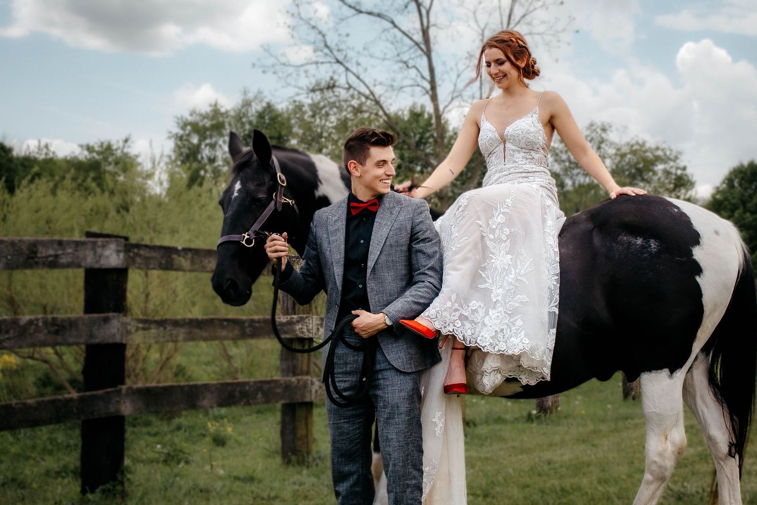 A couple poses playfully on a rustic wooden fence in a pastoral outdoor setting during their wedding photoshoot.