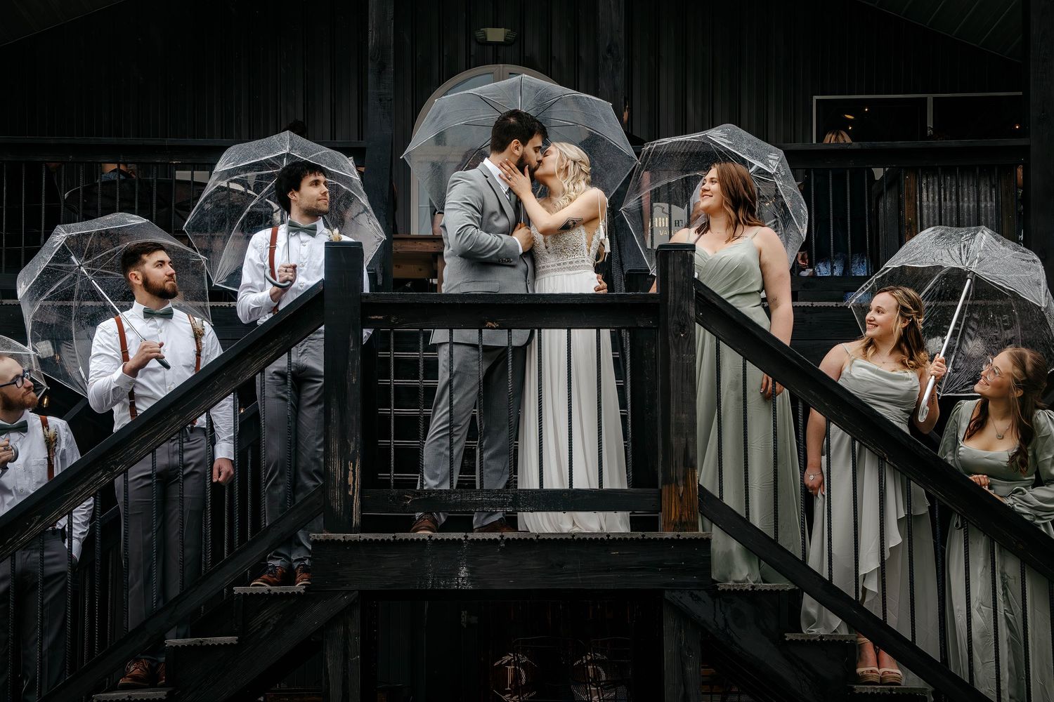 A romantic wedding couple poses with umbrellas on a dark industrial staircase in dramatic black and white photography.