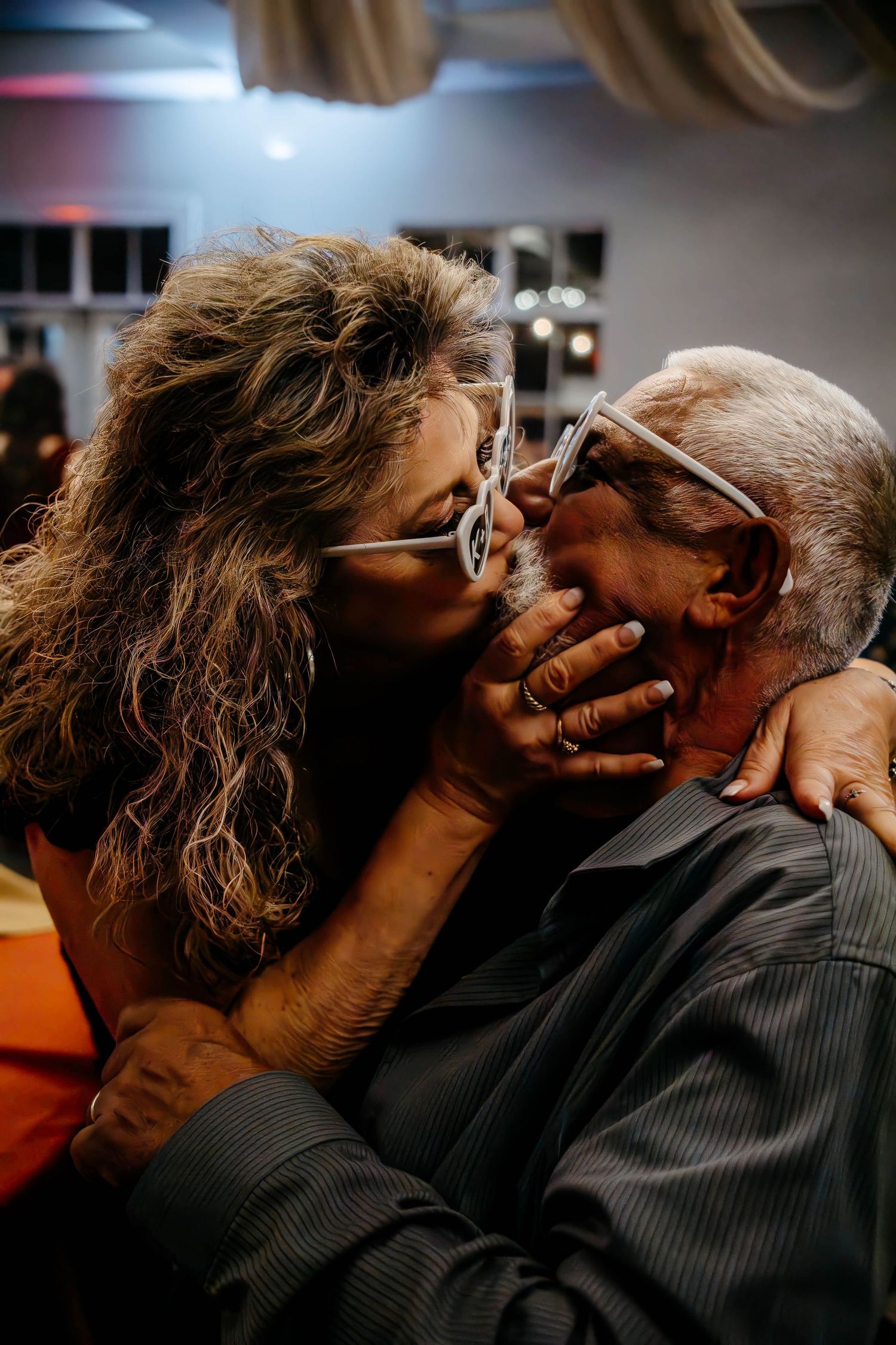 An older couple sharing a tender kiss, both wearing glasses, in a warmly lit indoor setting.