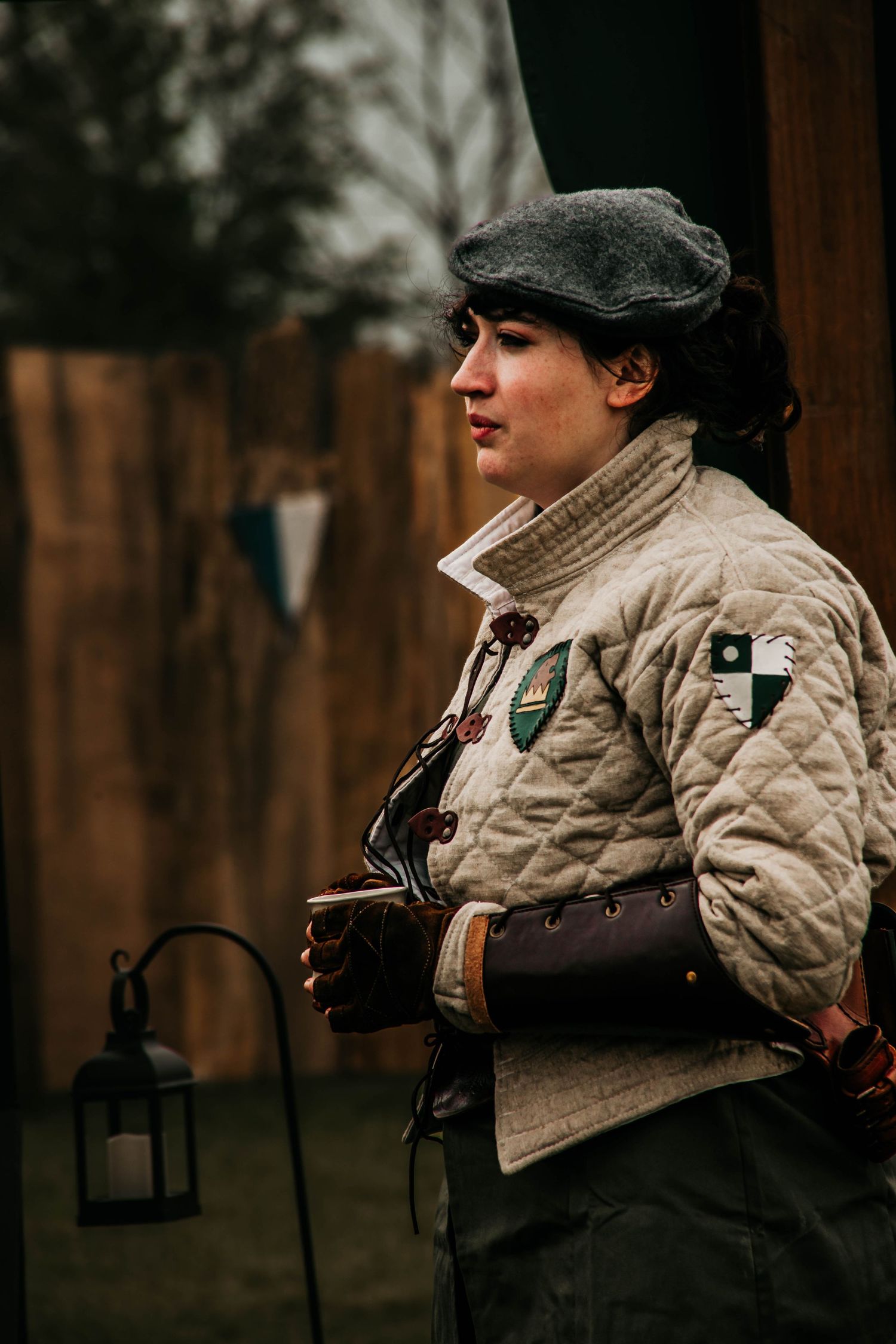 A person in a beige quilted jacket and dark hat stands outdoors against a blurred wooded background.