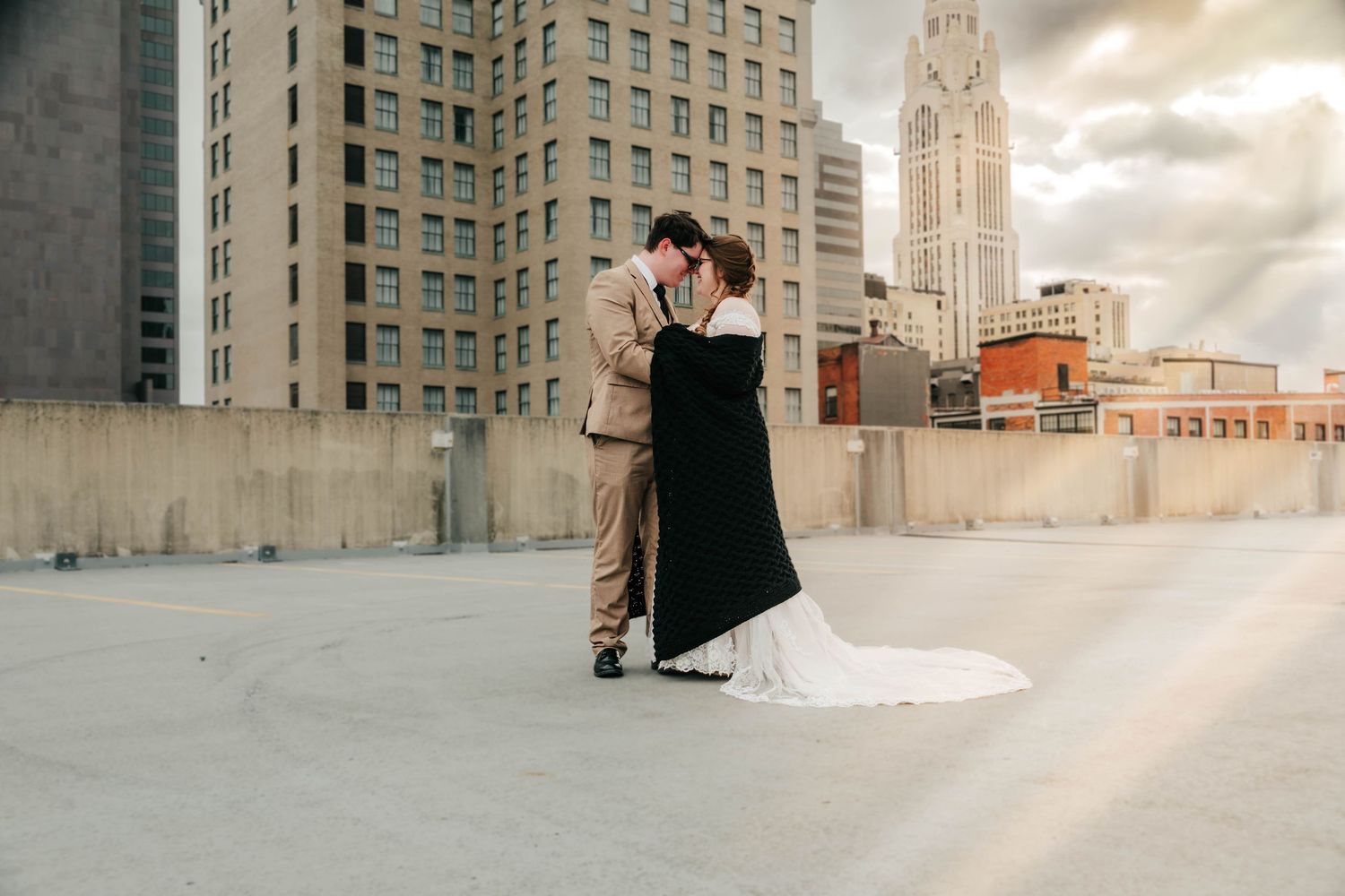 A couple embraces in front of an urban backdrop with tall buildings and dramatic cloudy sky at sunset.