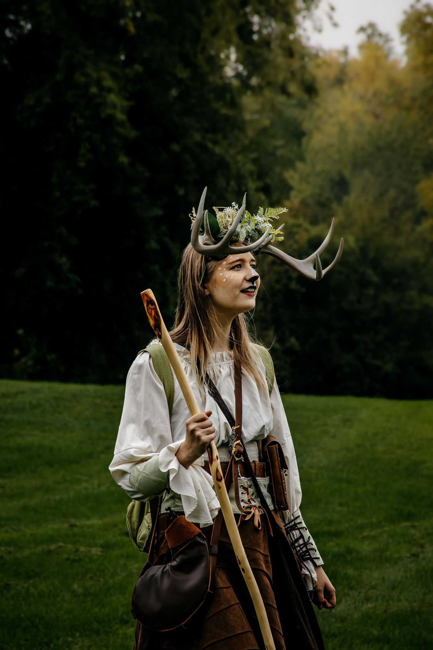 Woman in fantasy forest costume with antler headdress and bow, posing outdoors in a lush green park.