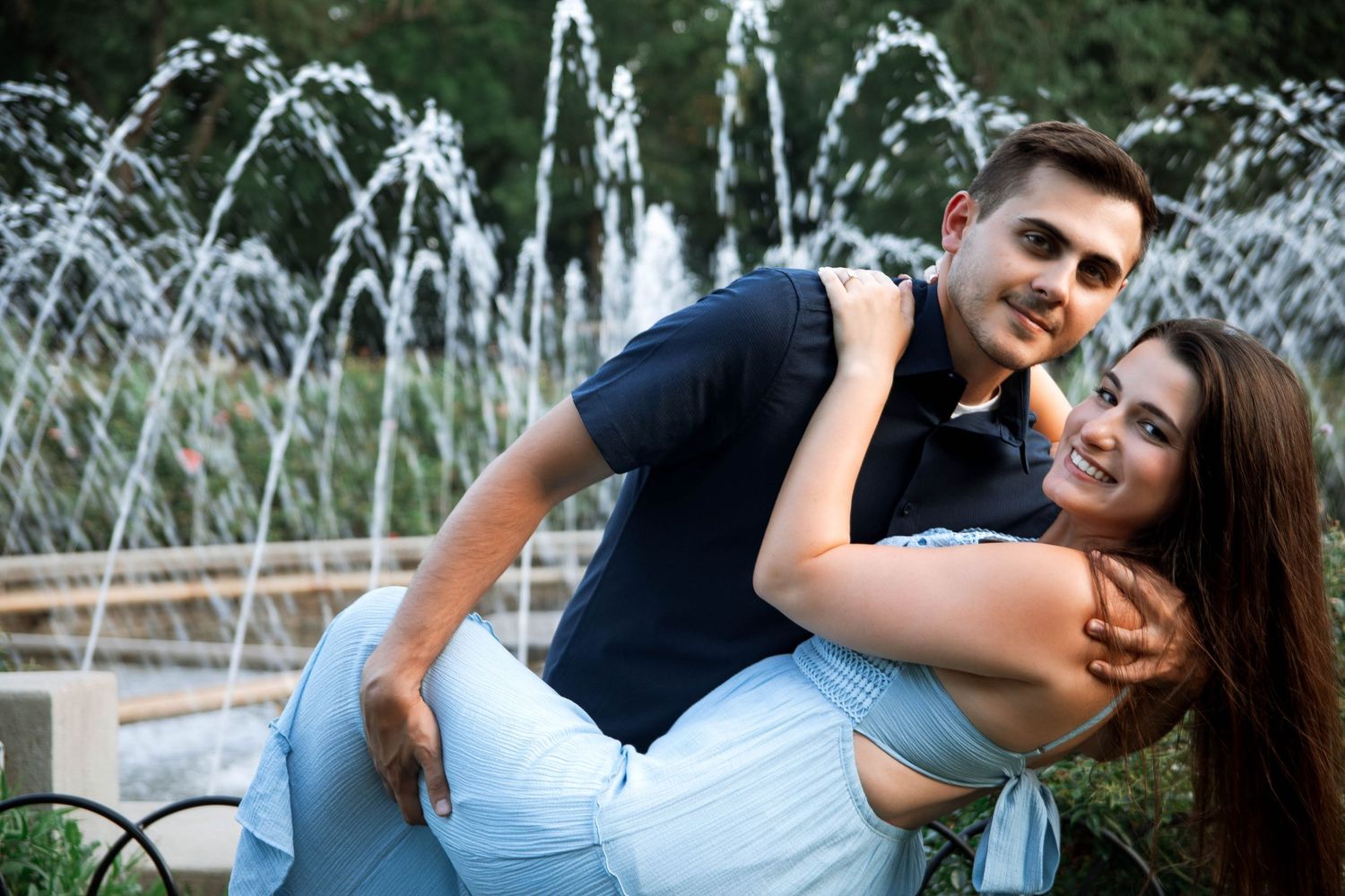 A couple embraces in front of a fountain during a romantic outdoor photoshoot in a garden.