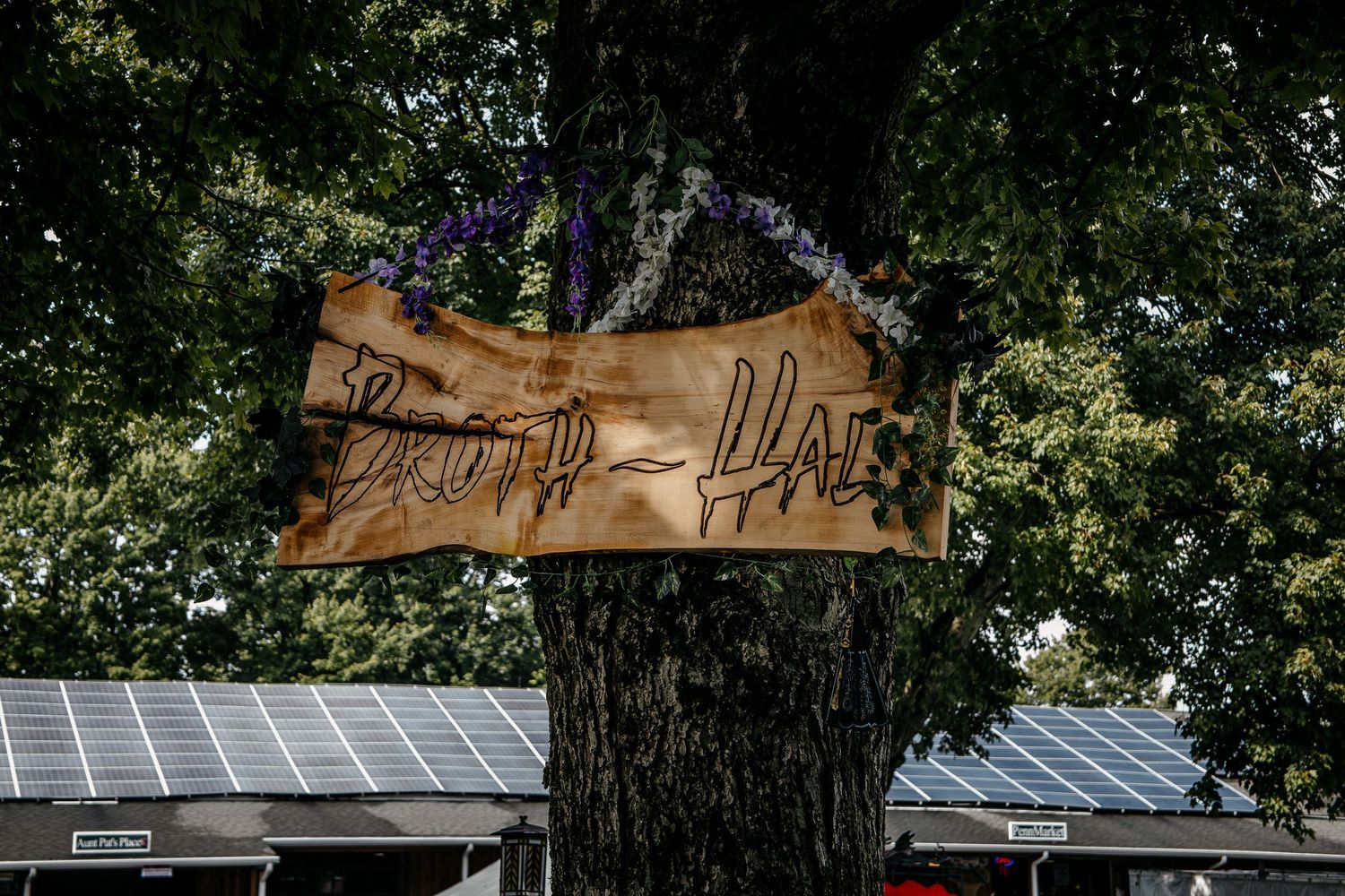 Wooden sign reading Brot-Haus hanging on a tree trunk outdoors with solar panels visible in background.