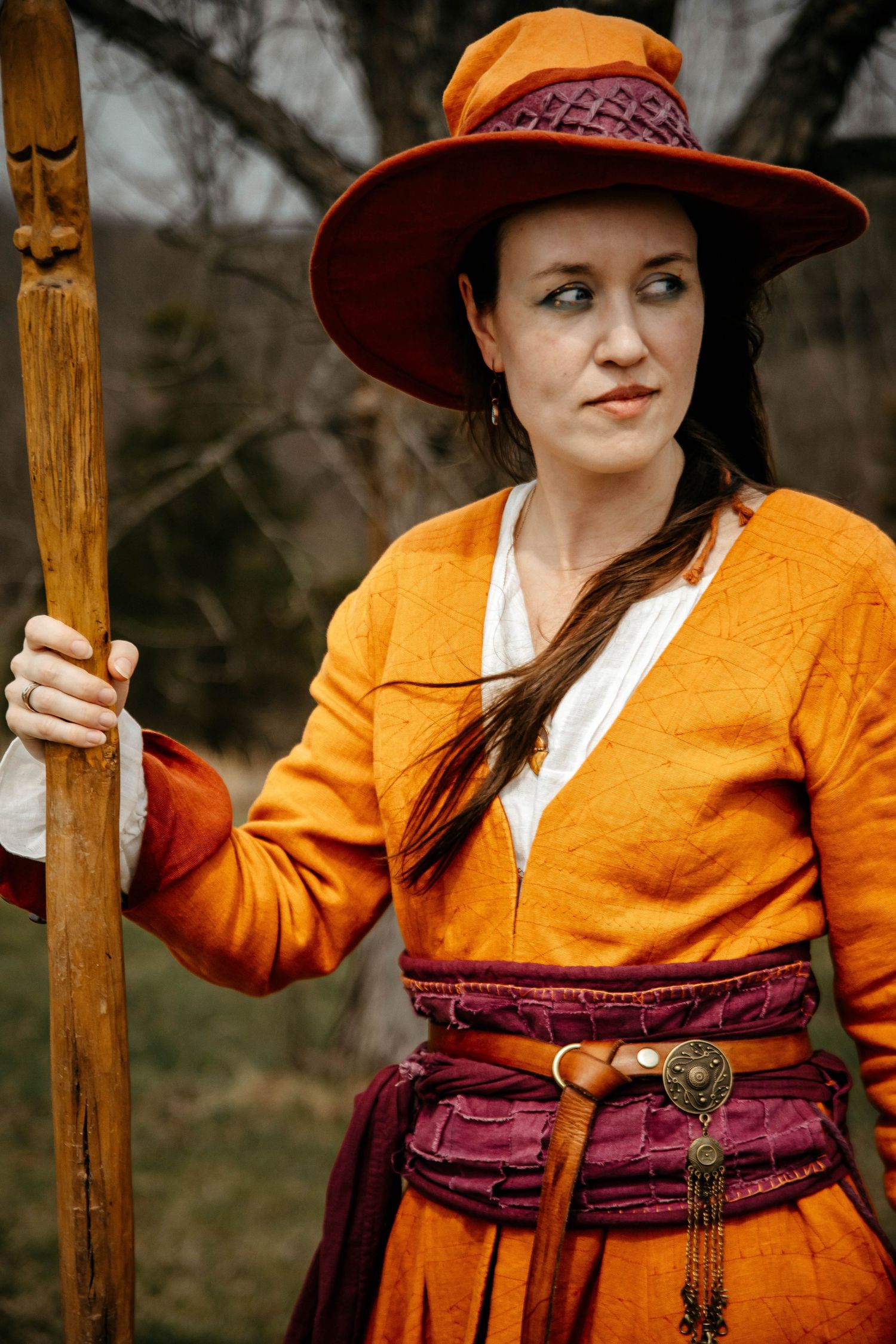 A person in a bright orange cardigan and burgundy skirt poses outdoors wearing a wide-brimmed hat and holding a wooden staff.