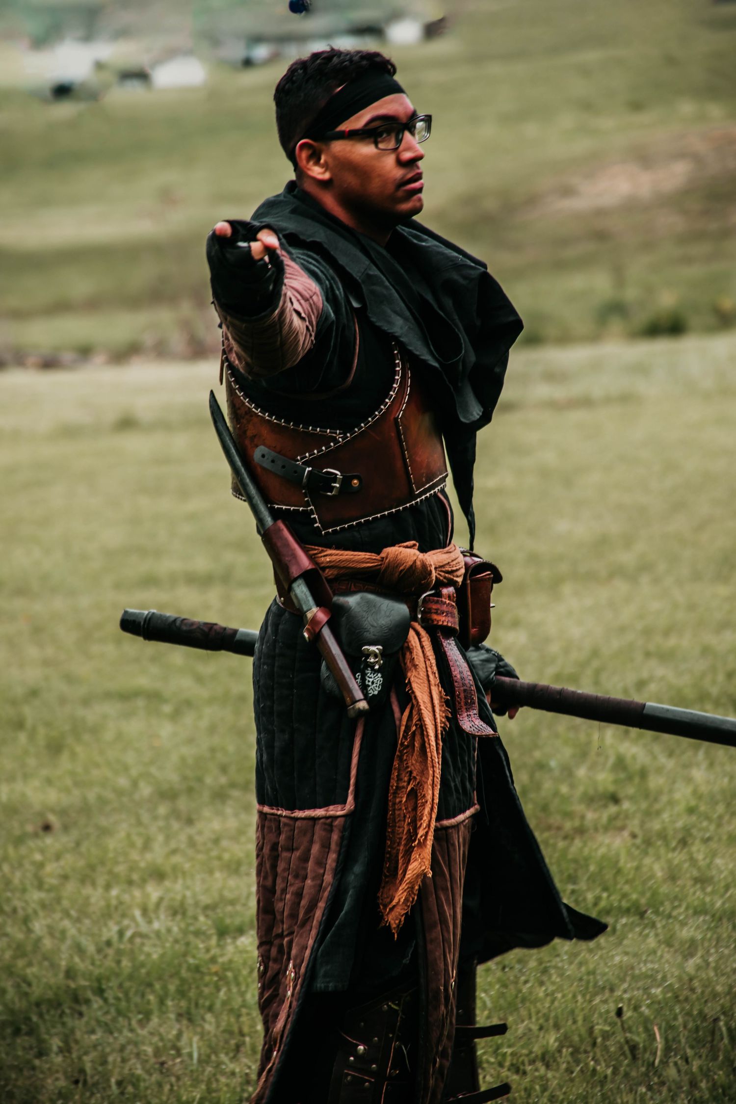 Person wearing traditional medieval costume with leather accessories stands in grassy field holding staff.