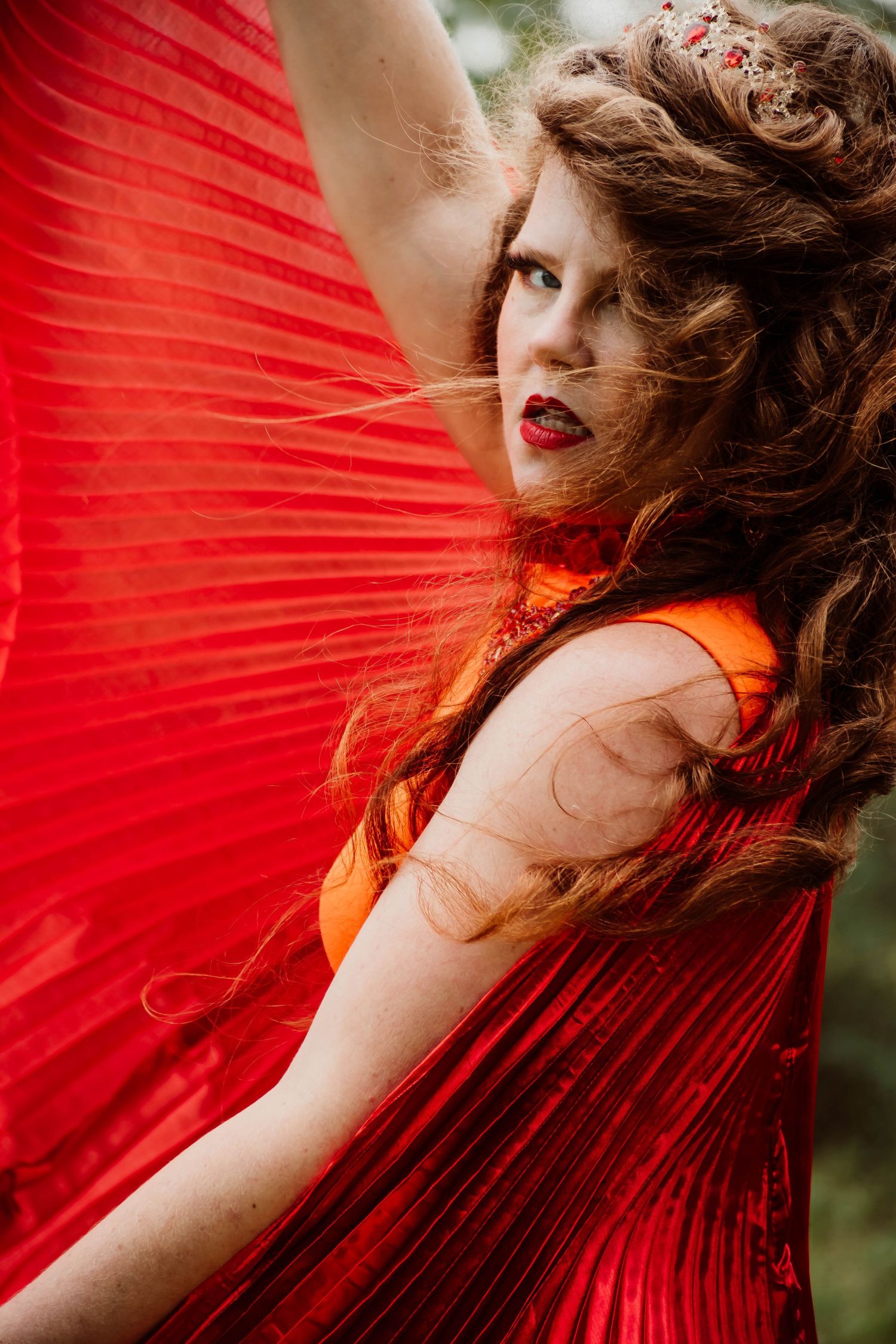 Woman in red dress holding a large red fan with curly hair and red lips, posing outdoors in a dramatic fashion.