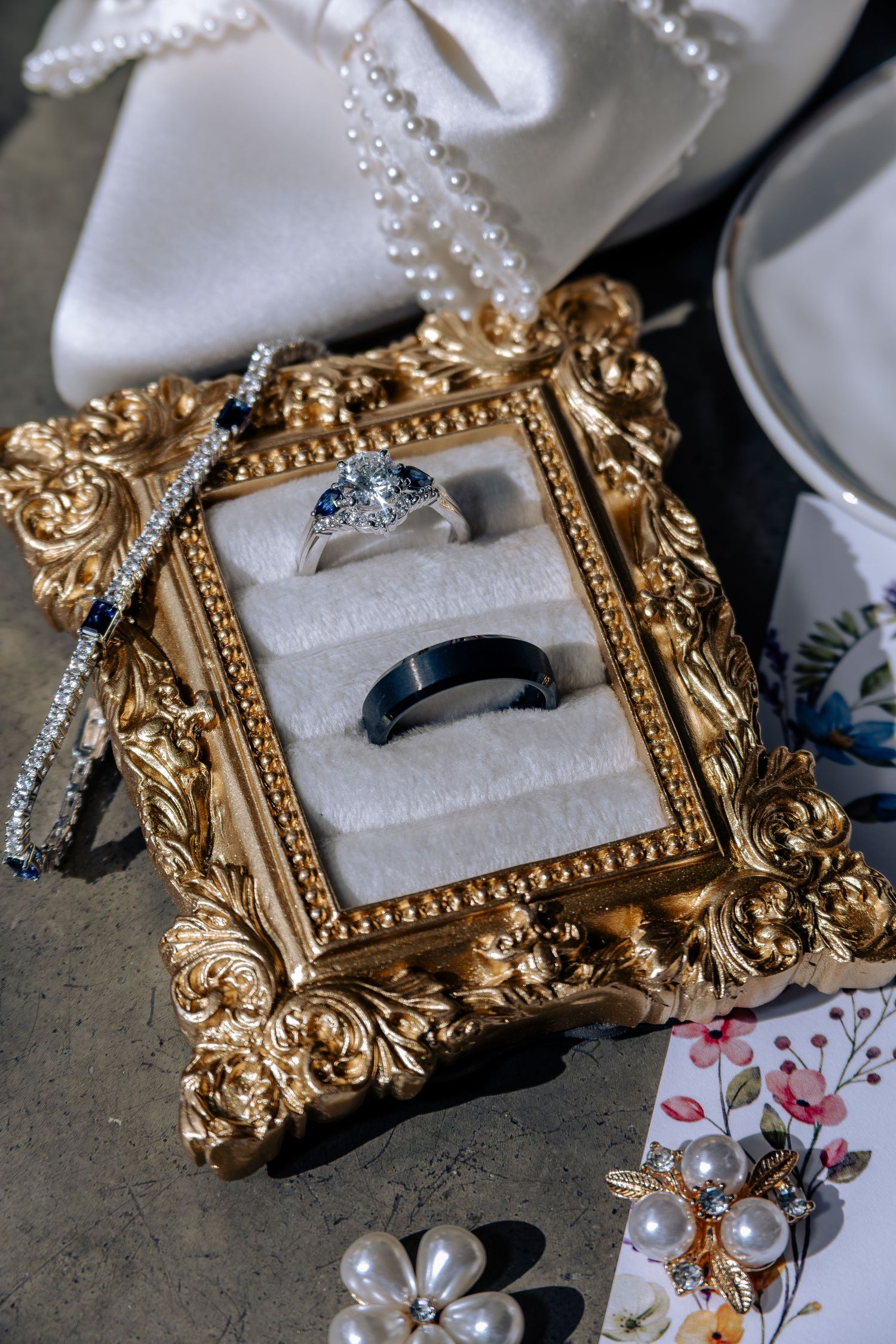 Elegant wedding rings displayed in ornate gold frame box with pearl necklace and floral card on marble surface.