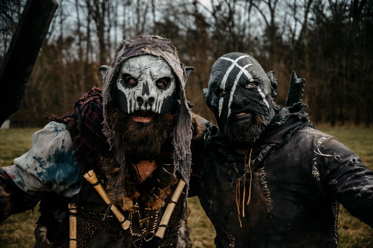 A person in a skull mask and dark clothing poses in a moody forest setting with weapons and dark atmospheric lighting.