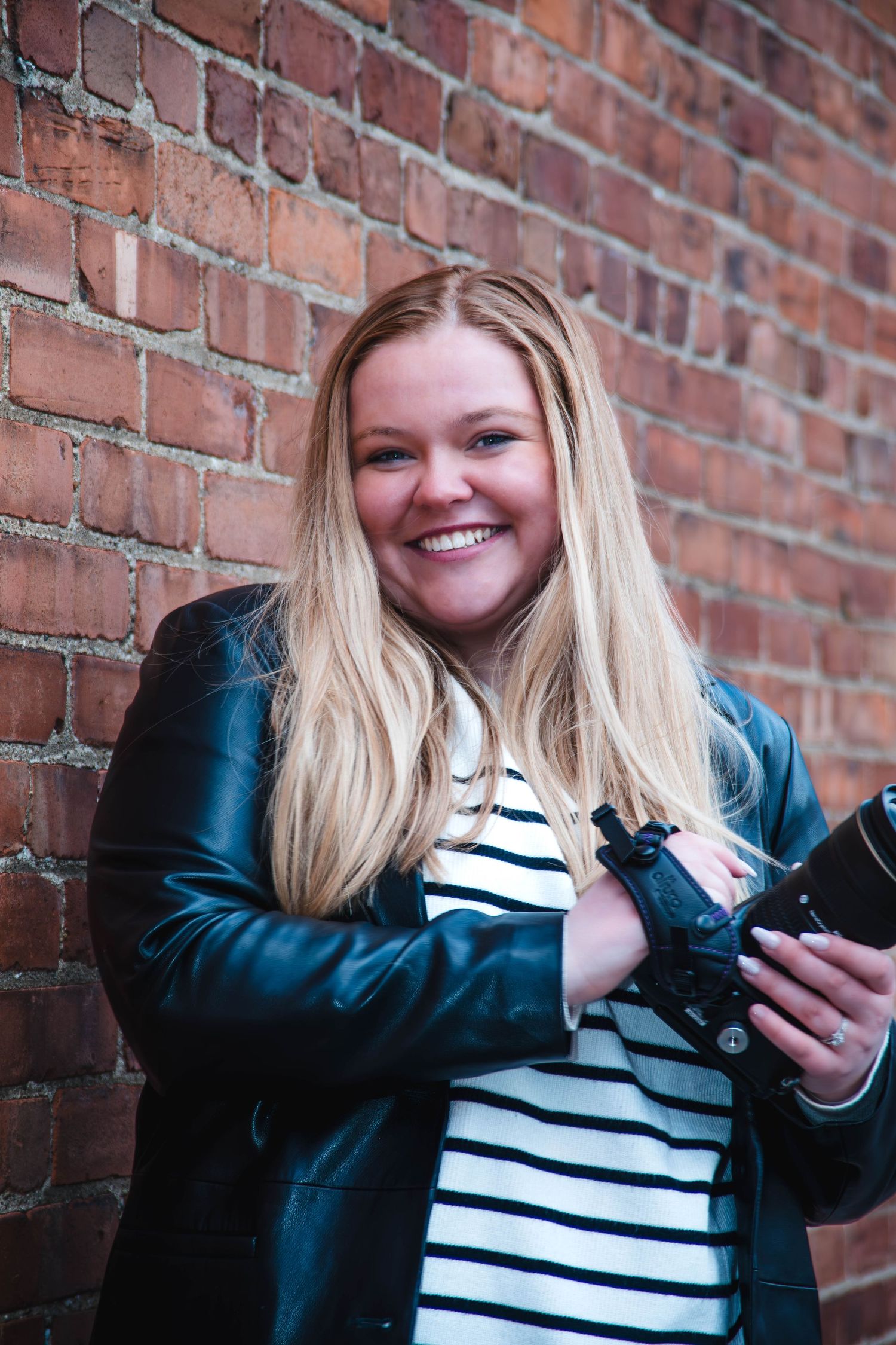 A person in a striped shirt and denim jacket poses against a brick wall while holding a phone.