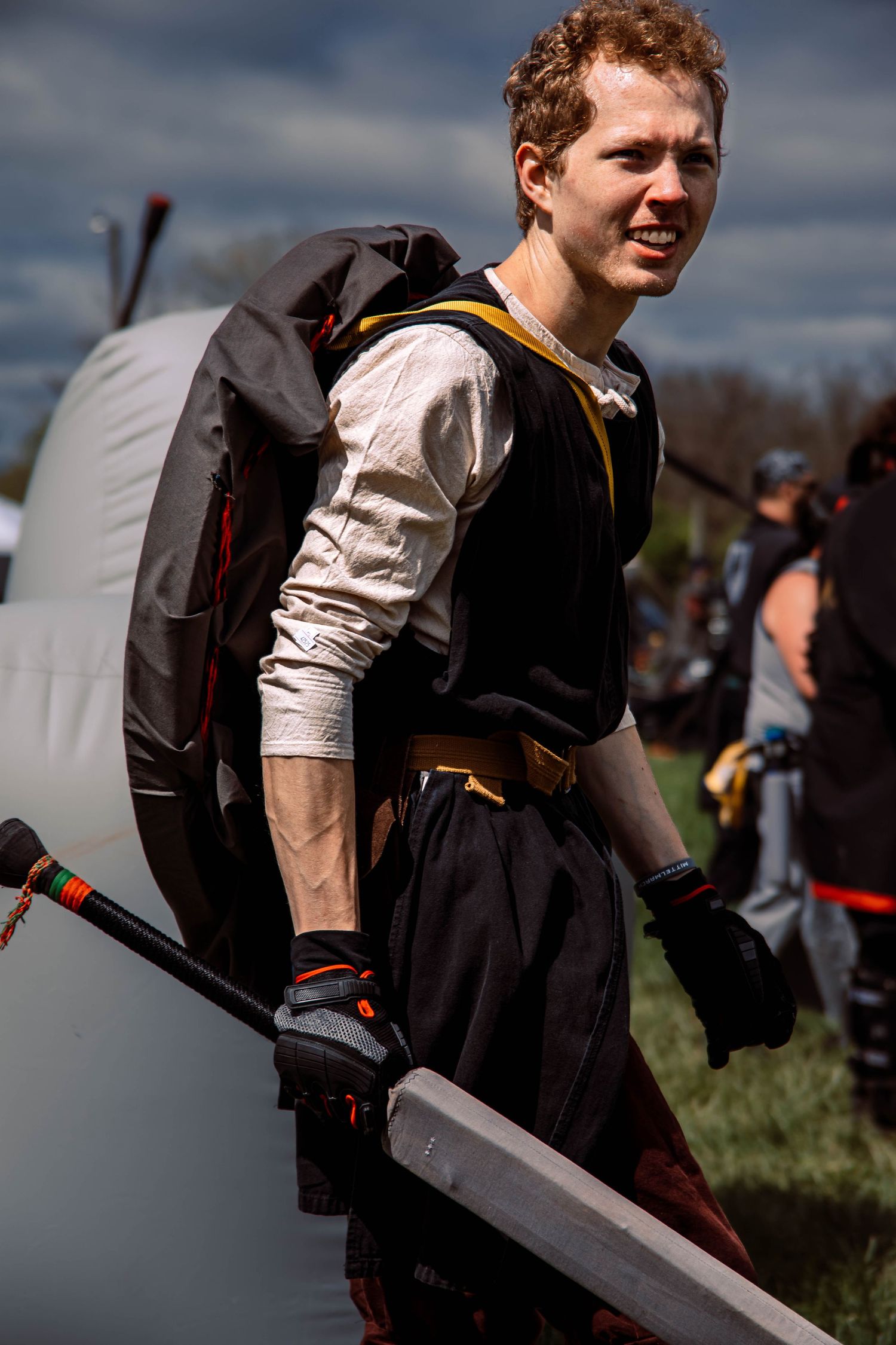 Medieval reenactor in black and white garments carries equipment during an outdoor event.