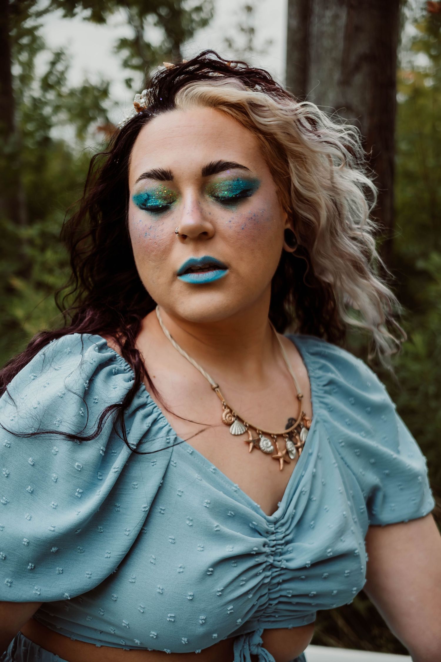 Fashion portrait in a teal vintage dress with dramatic blue makeup and forest backdrop.