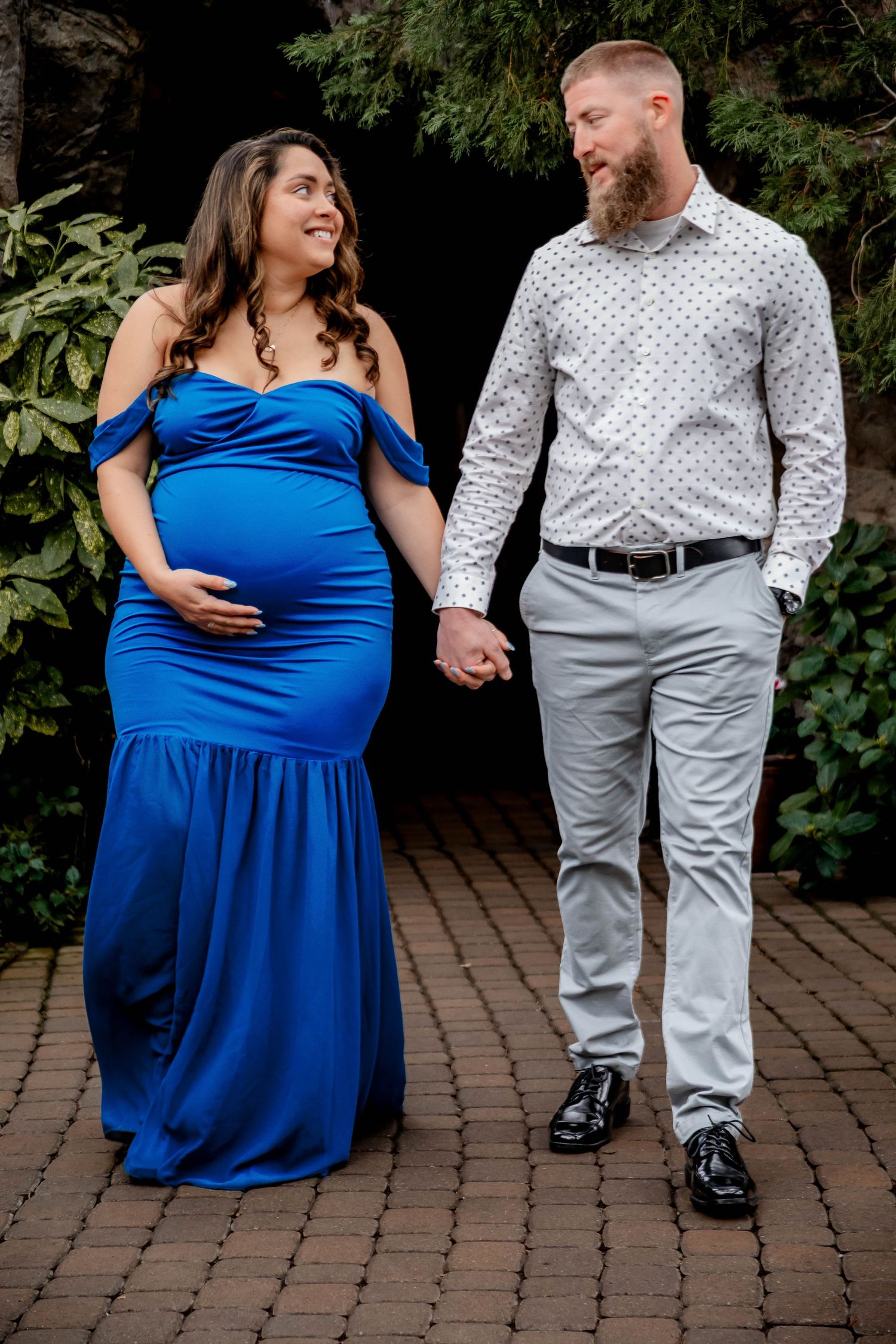 A couple holds hands while walking, with the woman wearing a stunning blue formal maternity gown.