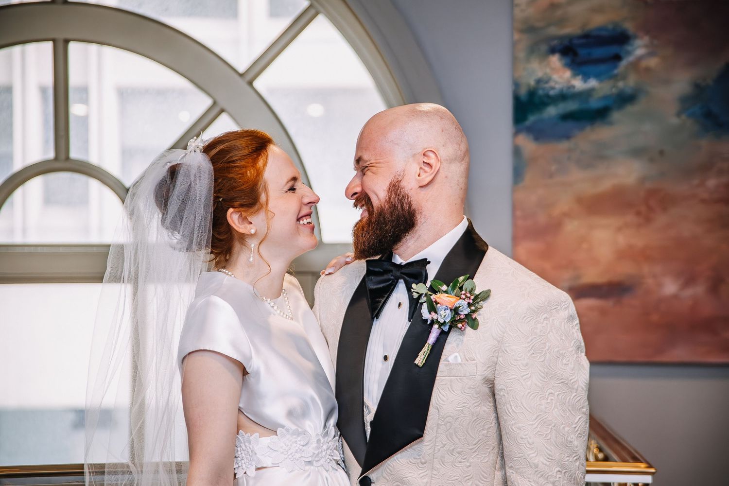 Bride and groom share a joyful moment, she in a white gown with veil, he in a cream tuxedo with boutonniere.