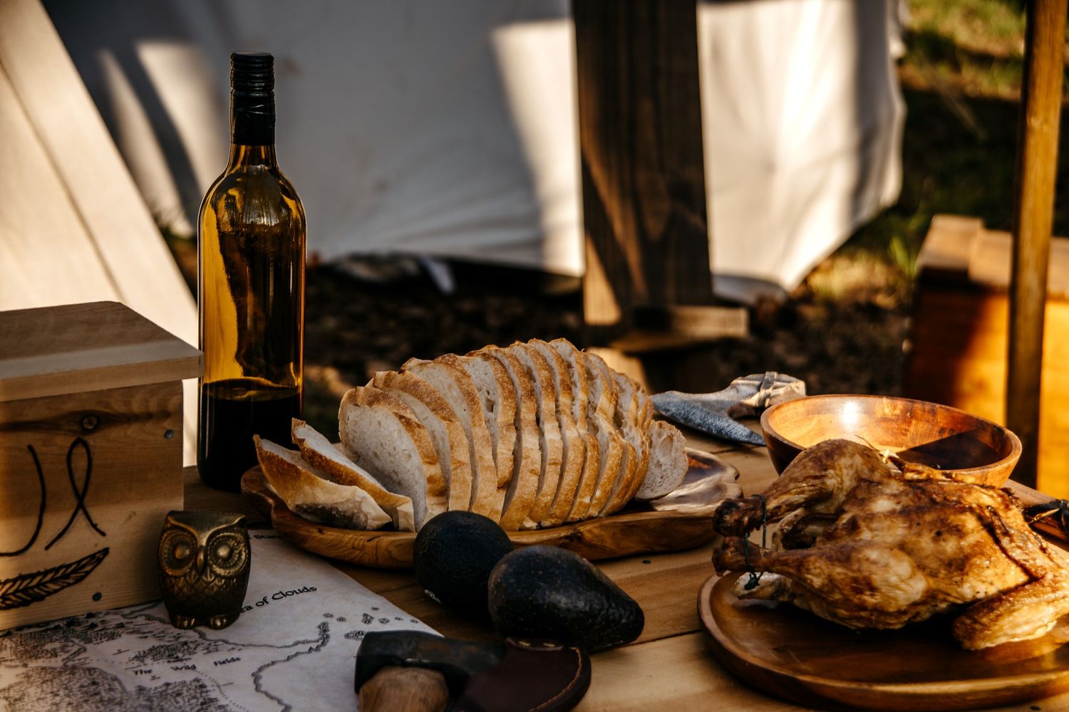 Rustic outdoor table with sliced bread, wine bottle, roasted meat, and owl figurine in warm golden light.