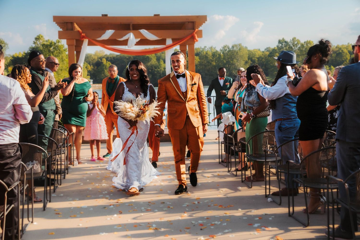 A joyful couple walks back down the aisle at an outdoor wedding ceremony under a wooden arch as guests celebrate.