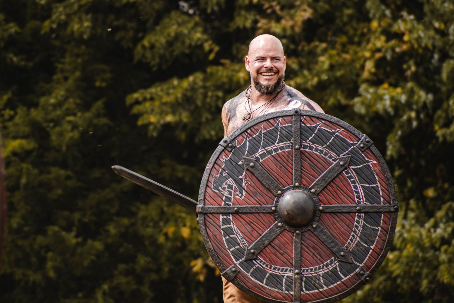 A smiling person in traditional Viking attire holds up a large decorated round shield against a green forest backdrop.