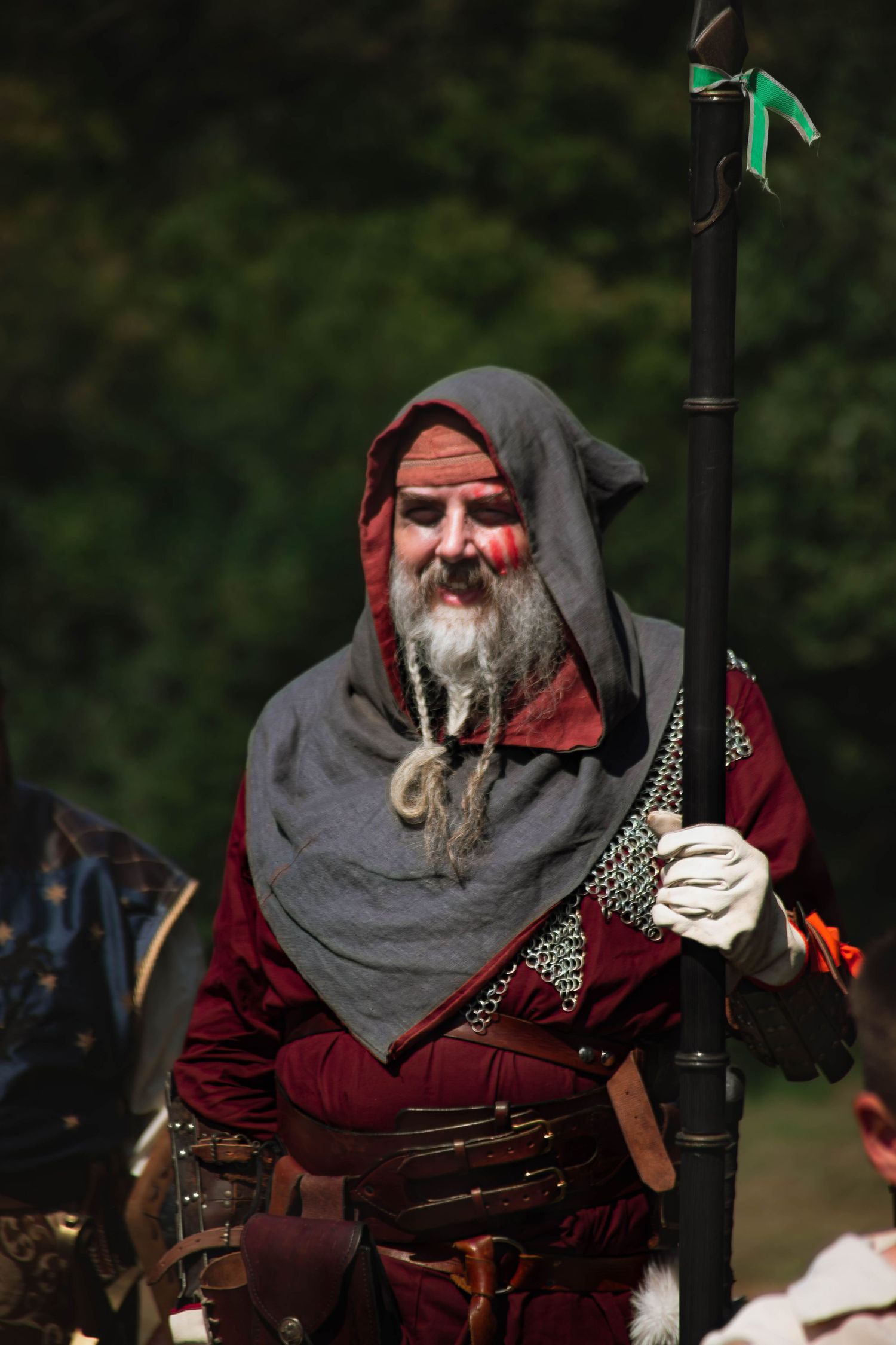 Medieval warrior in red tunic and gray hood with braided beard holds weapon during historical reenactment event.
