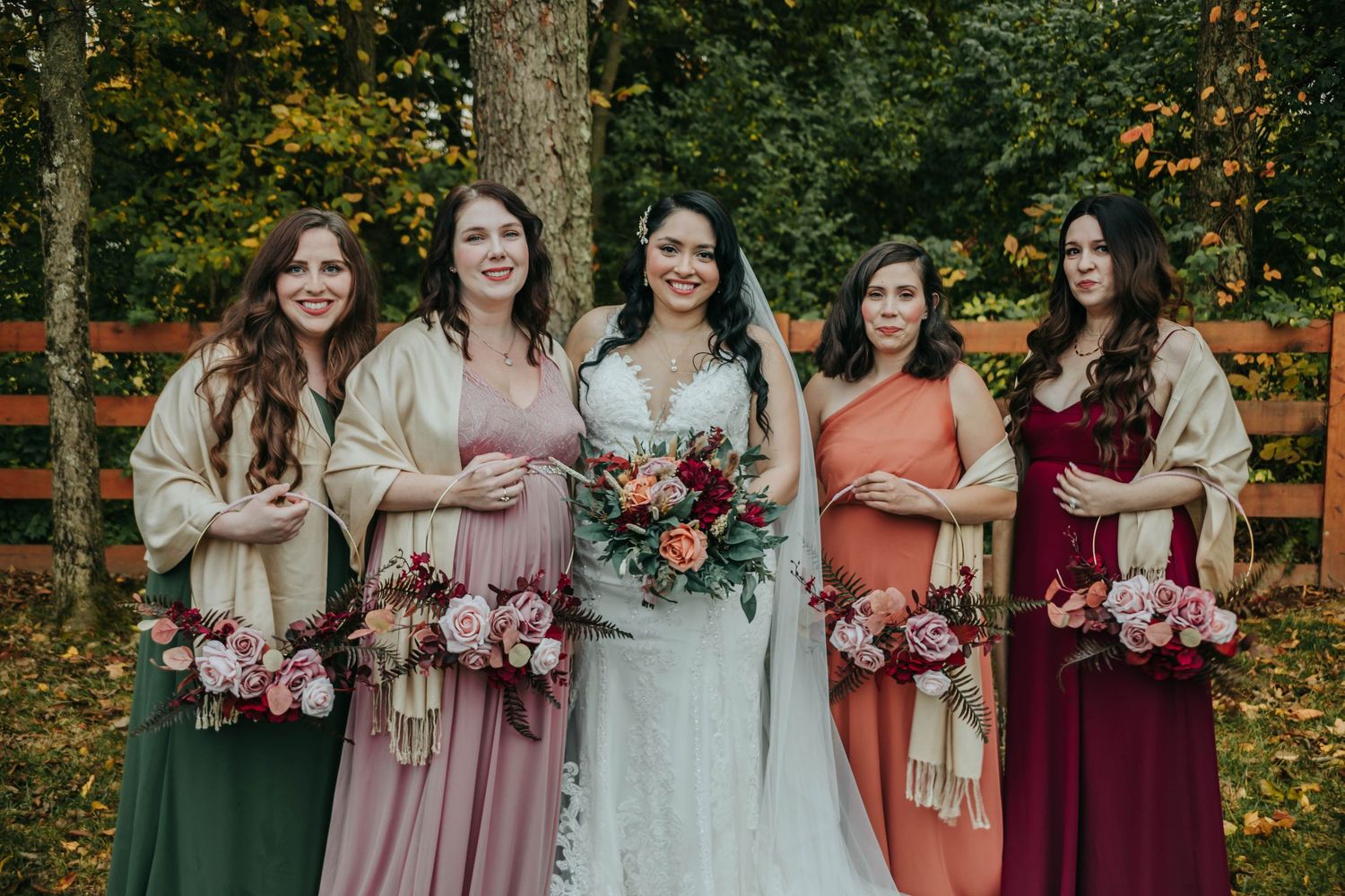 Bridal party wearing mix of burgundy, blush, and sage green dresses poses together in autumn forest setting.