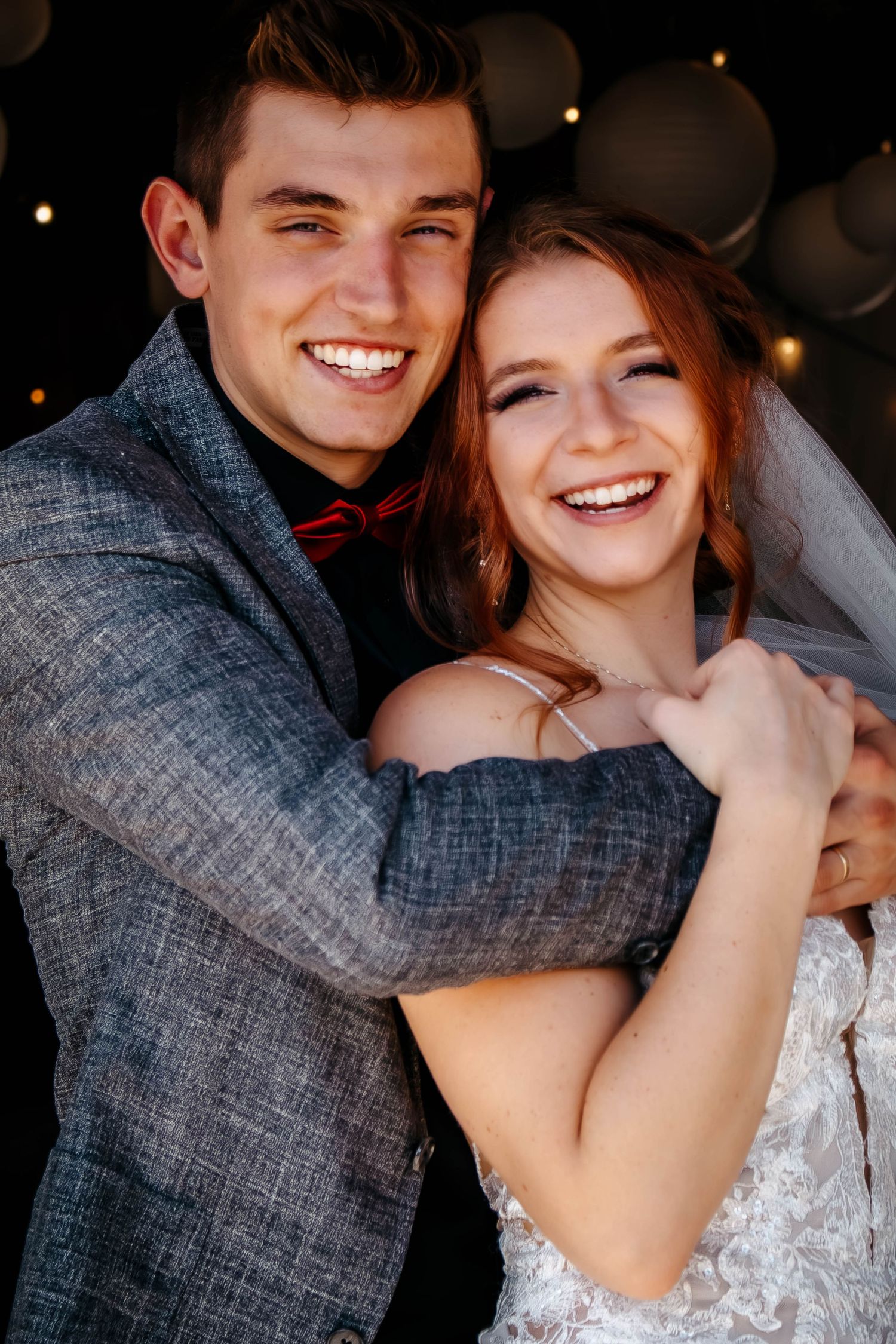 A joyful couple embraces on their wedding day, with the bride in white and groom in gray suit with red bowtie.