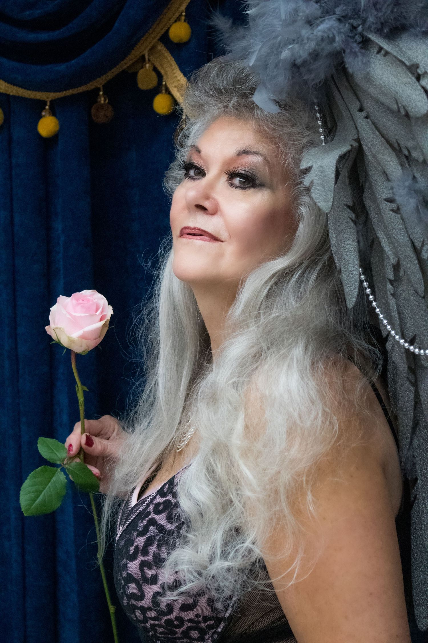 Elegant portrait with long silver hair, feather headpiece and pink rose against blue curtain backdrop with gold tassels.