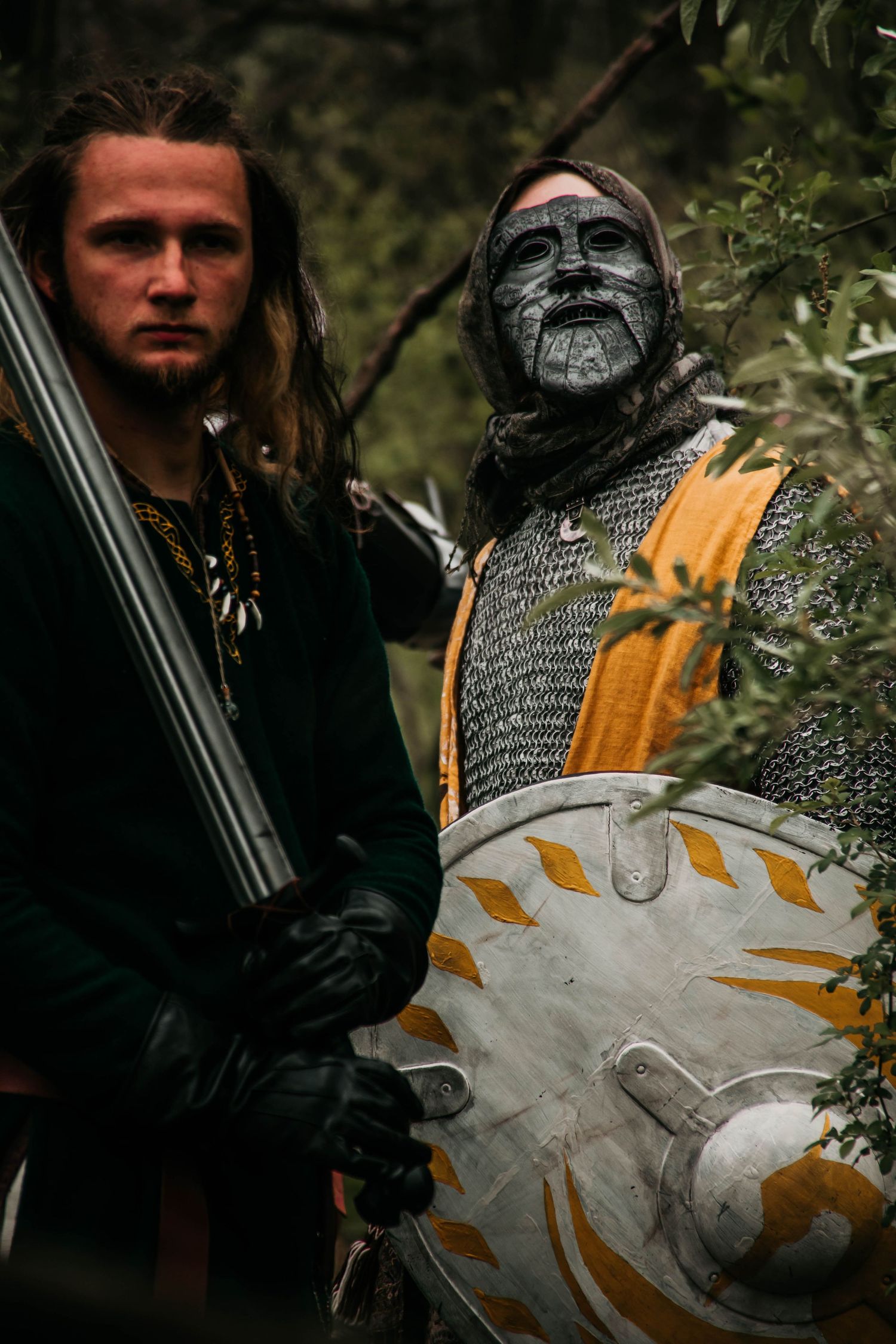Medieval knight in chainmail armor and yellow cloak examines ornate shield with sun emblem in forest setting.