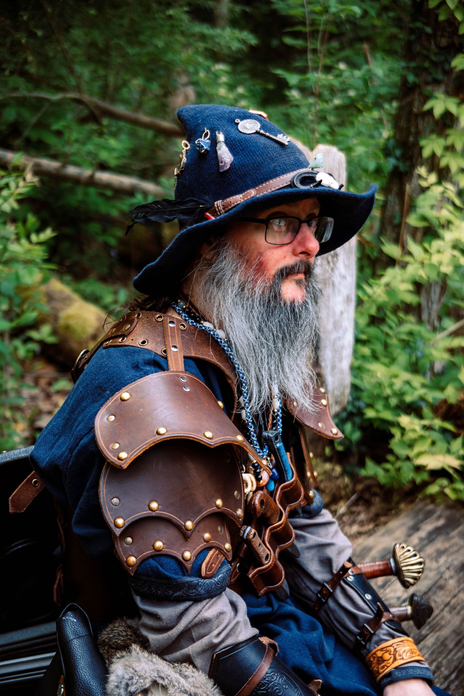 A person in steampunk attire wearing a decorated leather armor and blue hat with glasses in a forest setting.