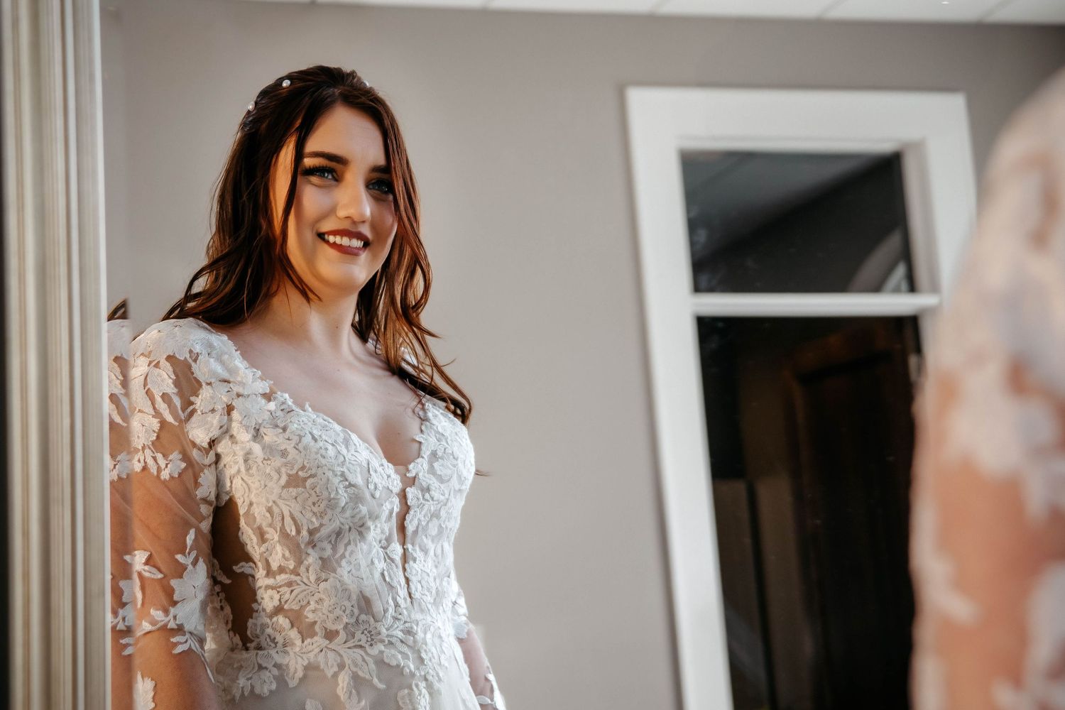A bride smiles while wearing an elegant lace wedding dress with sheer sleeves.