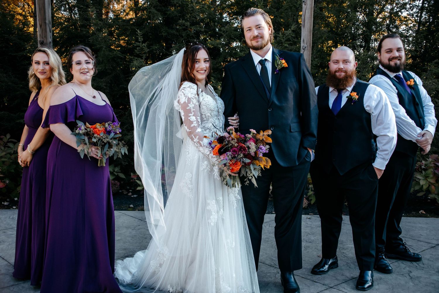 Wedding party members in purple dresses and dark suits gather for formal photos outdoors.