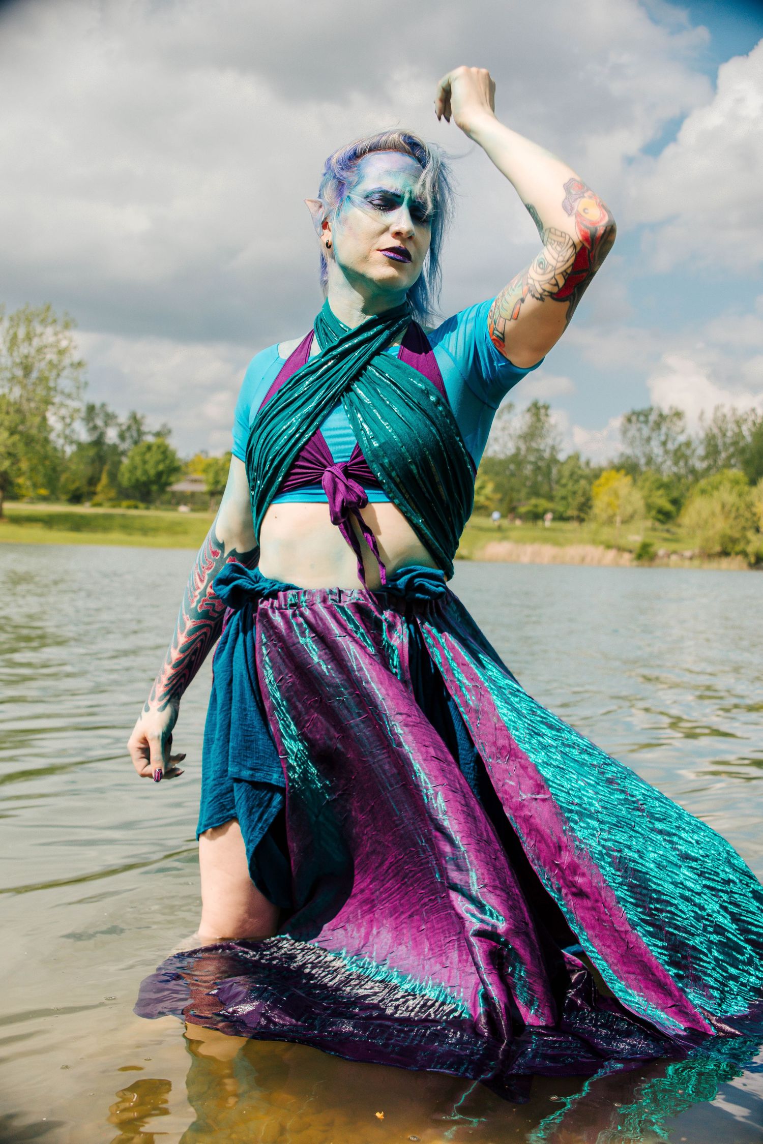 Person in teal and purple costume with blue hair standing in water, wearing sequined skirt and dramatic fantasy makeup.