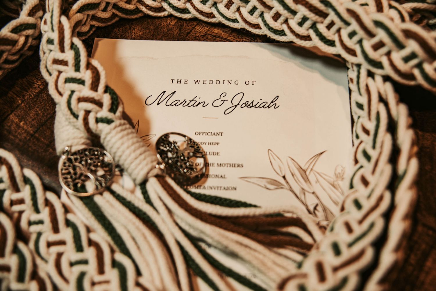 Wedding invitation for Martin and Josiah displayed on a wooden surface with decorative Celtic knot rope and ornate silver tassels.