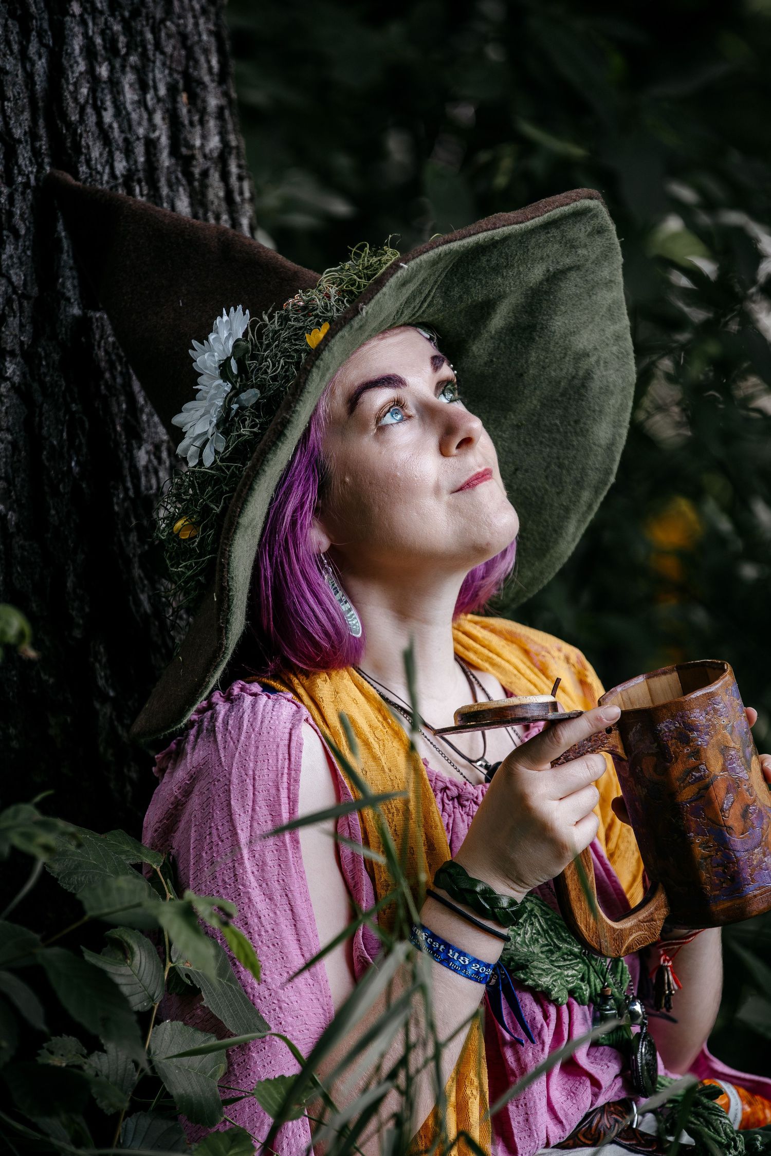 A woman in a green witch hat with flowers and pink hair holds a wooden mug while sitting by a tree outdoors.