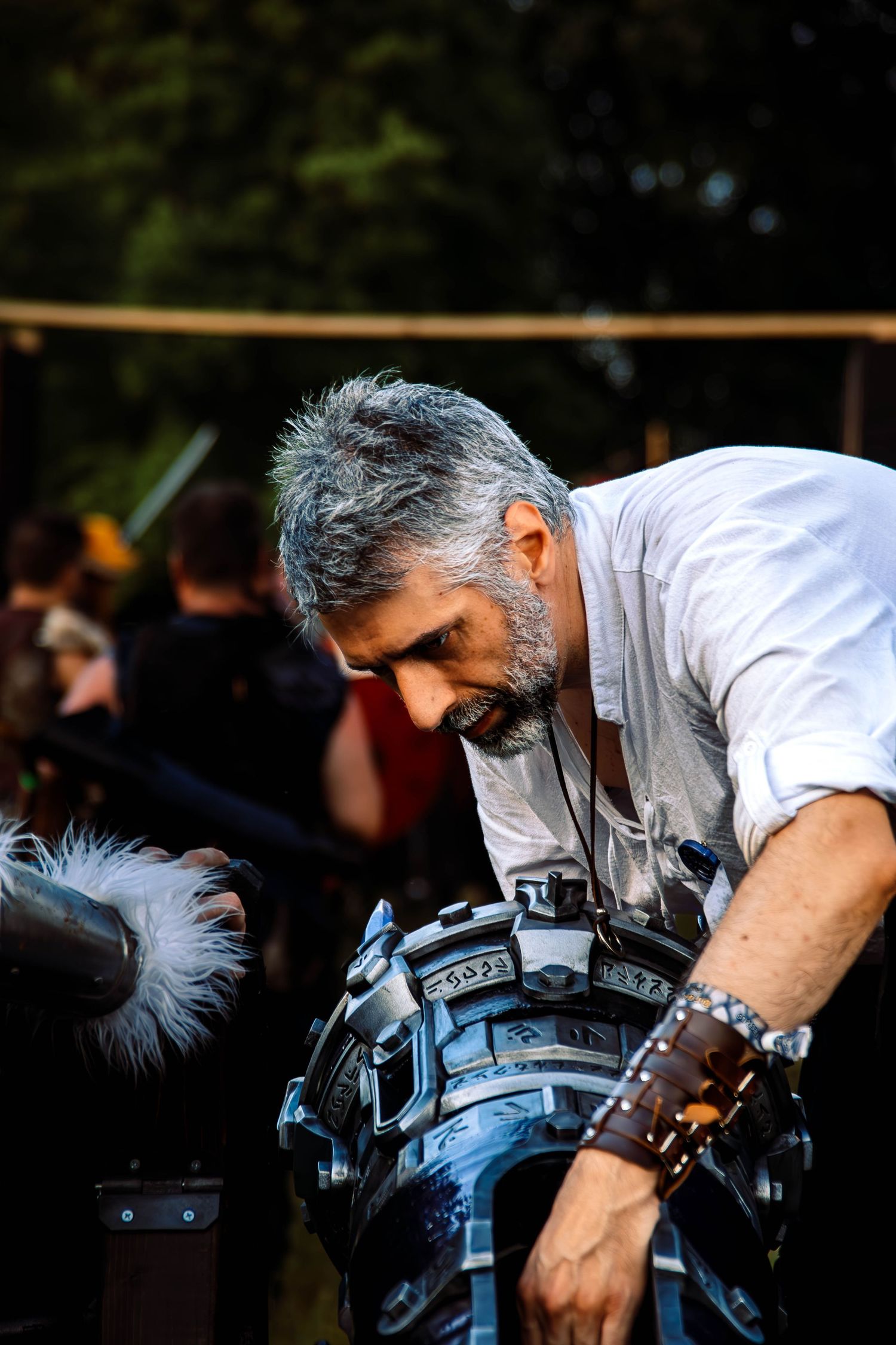 Gray-haired bearded man in white shirt examining elaborate steampunk armor at outdoor medieval festival.