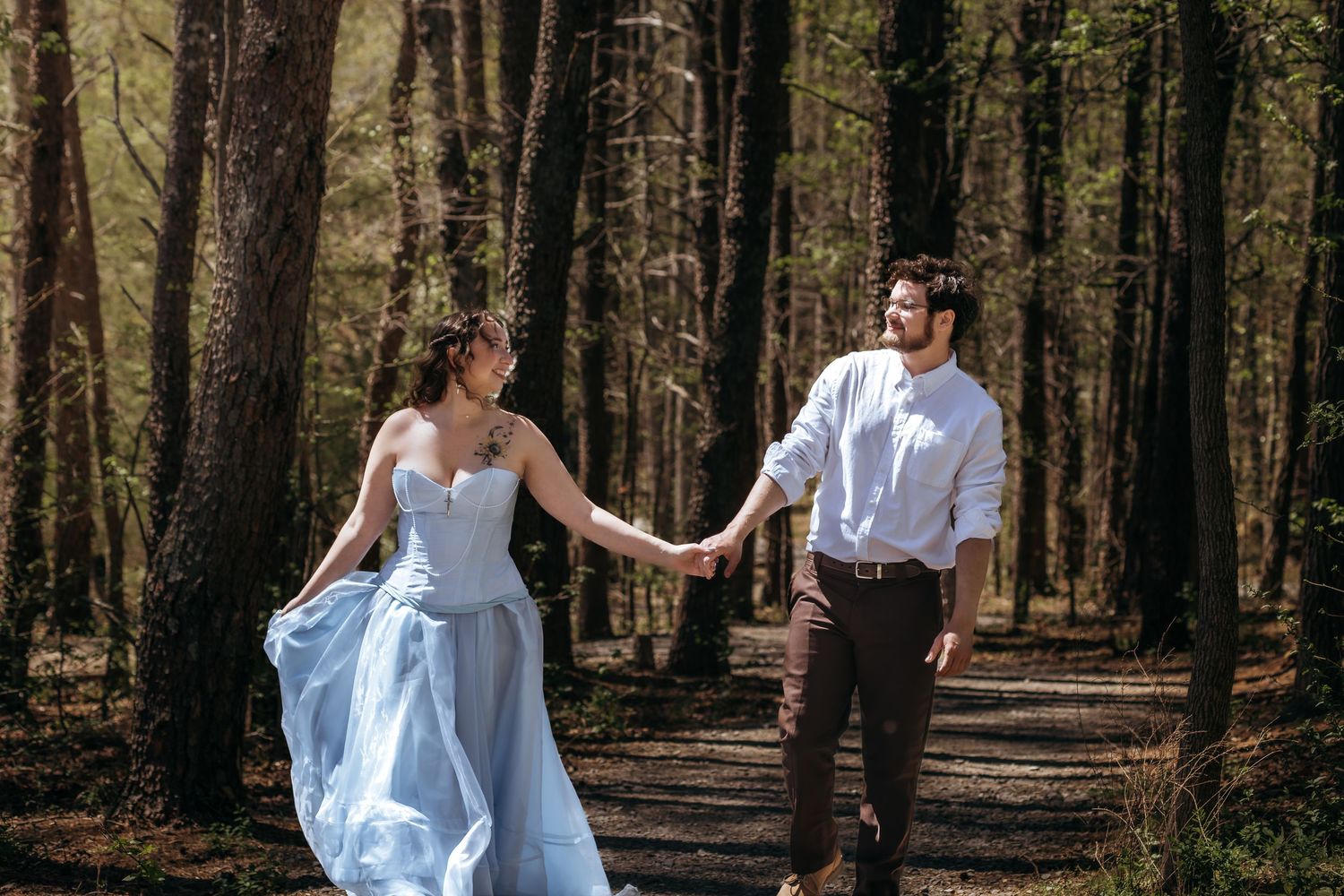 Couple holding hands walking through a sunlit forest path, woman in flowing blue gown, man in white shirt.