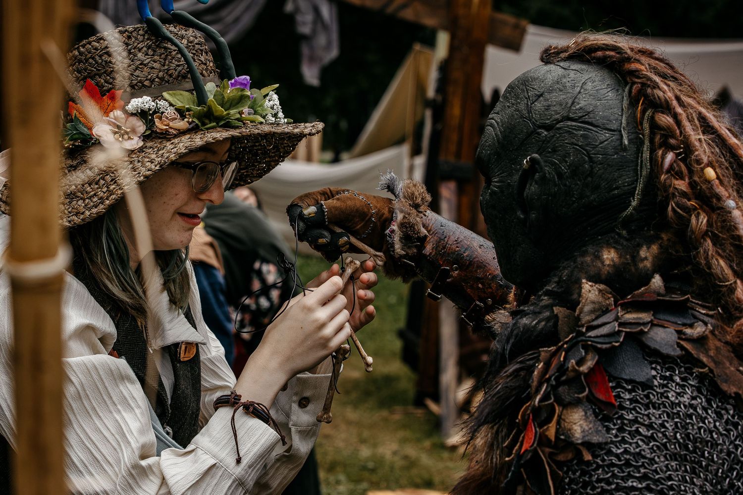 Two costumed performers at a medieval fair, one in a floral hat examining an antique pistol held by a dark armored creature.