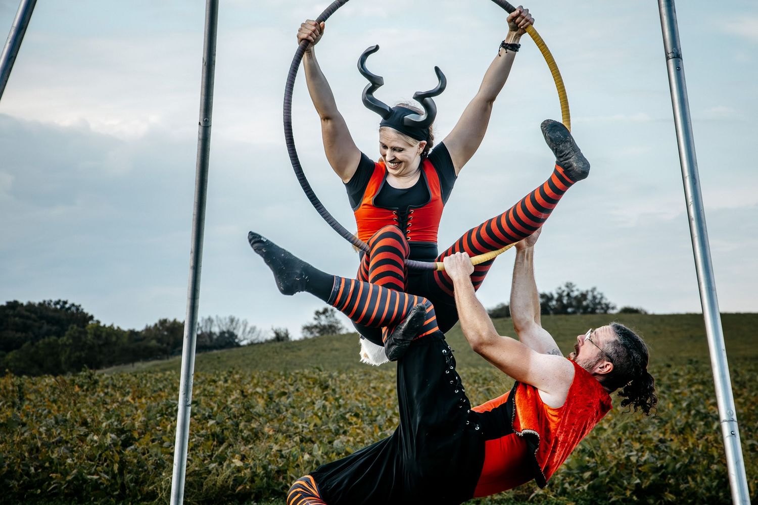Two acrobats in Halloween costumes perform aerial hoop tricks outdoors, one wearing devil horns and striped socks.