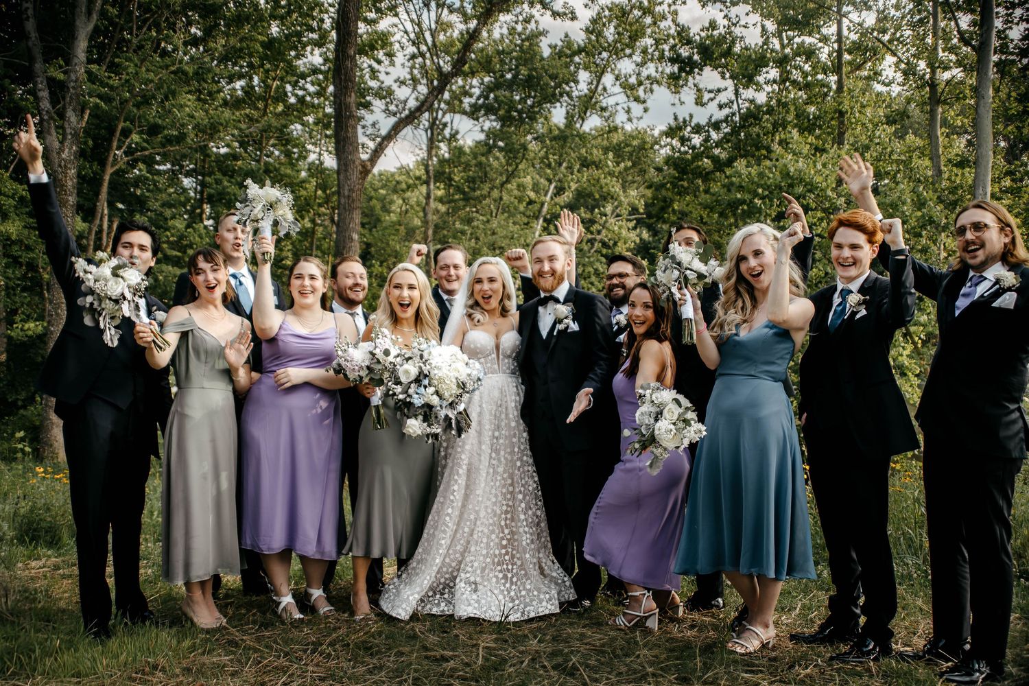 Wedding party celebrating outdoors in forest, bridesmaids in purple and grey dresses, groomsmen in suits, bride and groom in center.