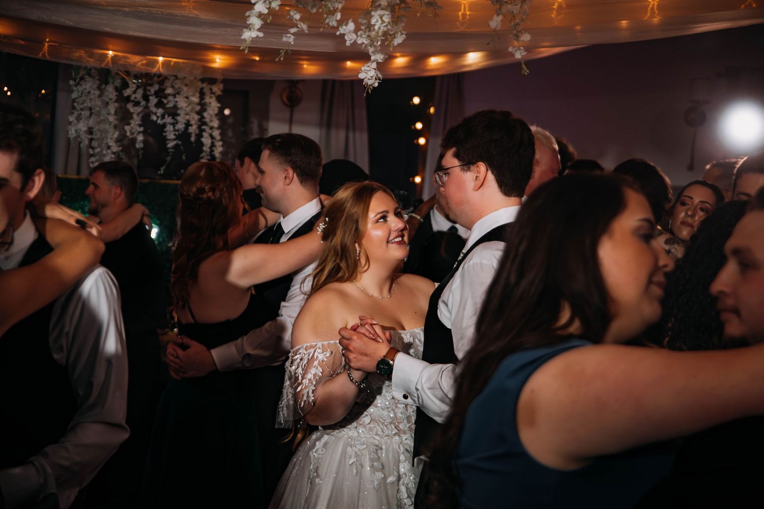 Bride and groom dancing joyfully with guests at a wedding reception under warm string lights.