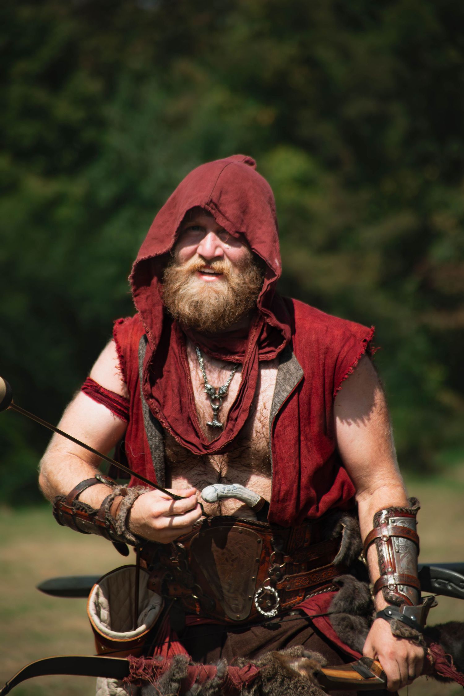 A warrior in red medieval garb and leather armor rides a horse during a reenactment event.