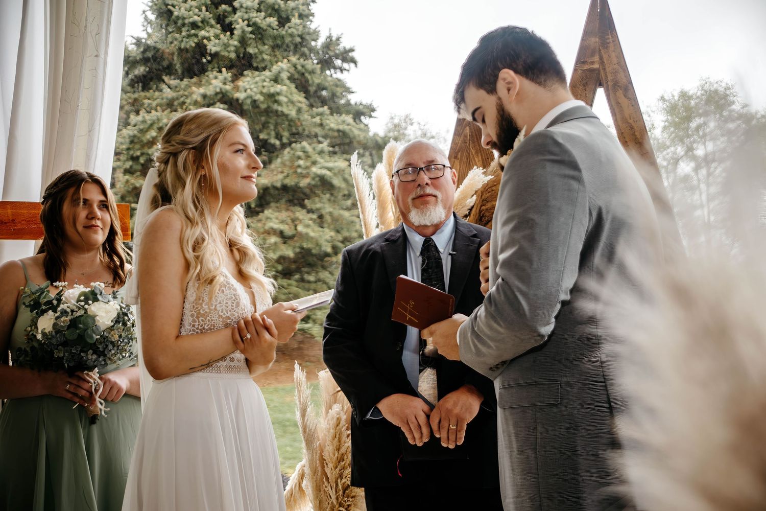 A wedding ceremony takes place under a wooden arch with a couple exchanging vows in front of an officiant.