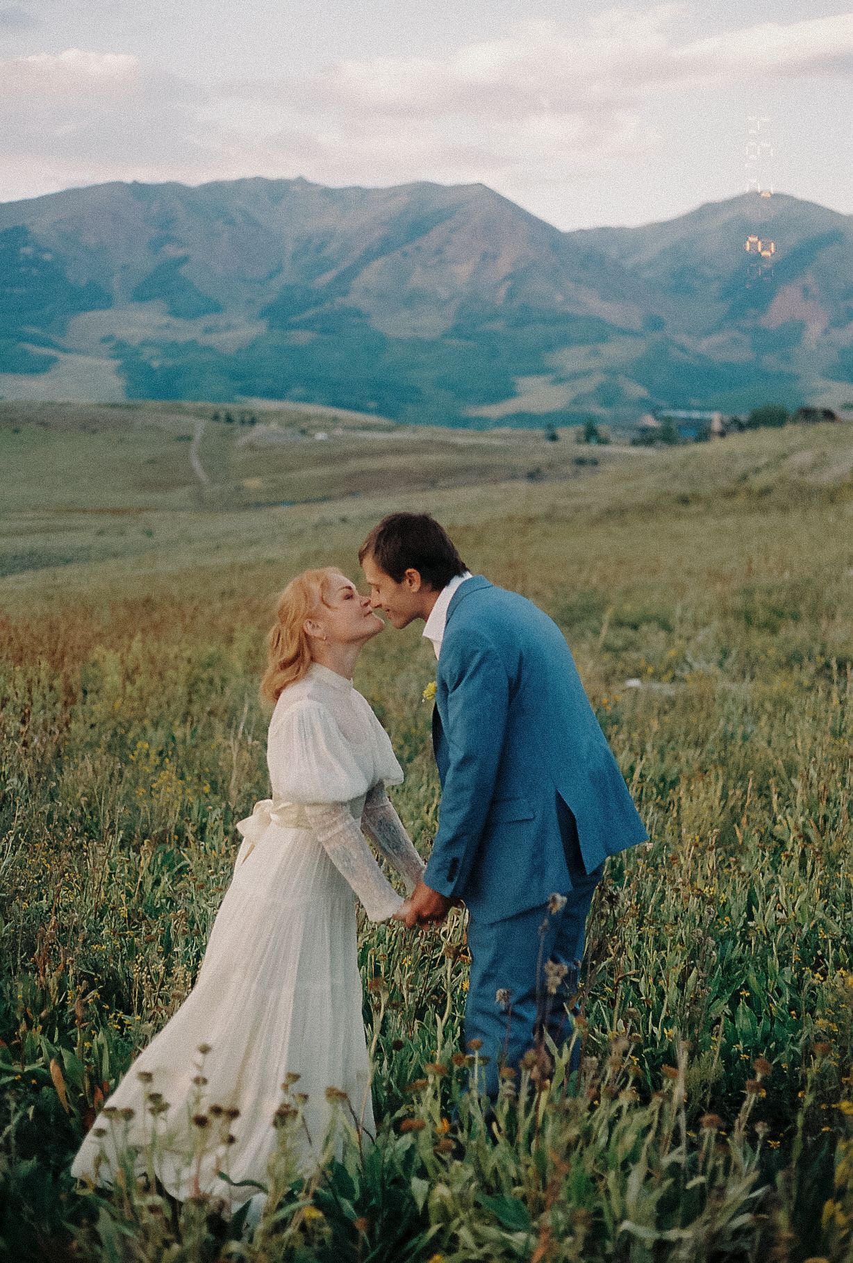 Bride and groom sharing romantic kiss in wildflower meadow with sweeping mountain views at dusk.
